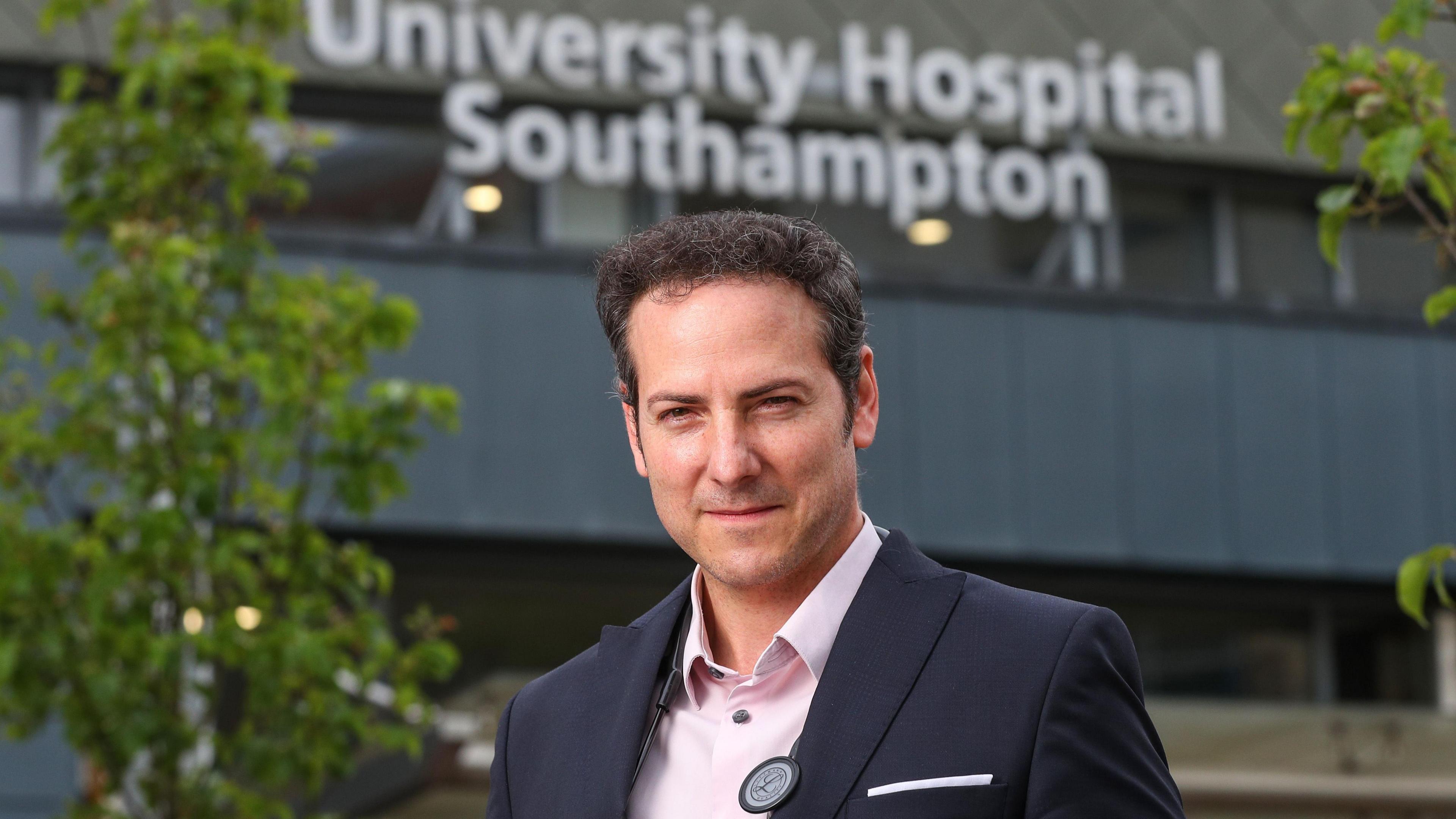 Dr Hugo stands in front of the University Southampton Hospital, which is in the background. He looks at the camera with his eyes narrowed. He is wearing a navy blue jacket and a pink shirt. There is a tree with green leaves between him and the hospital. 