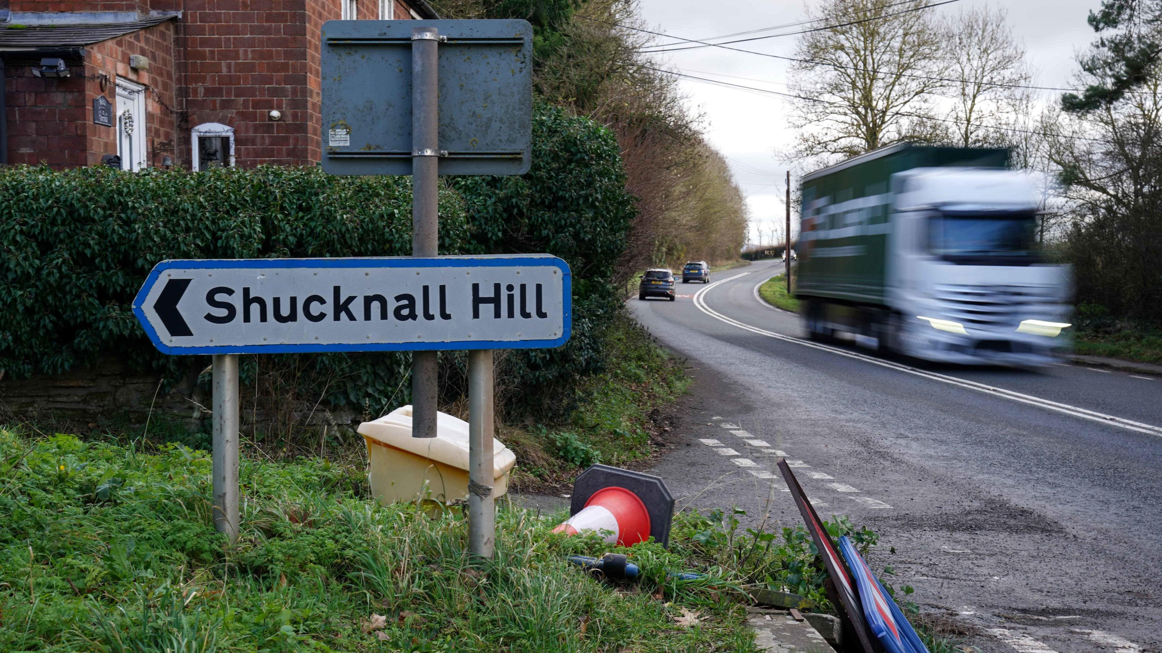 A road sign which shows Shucknal Hill. There is a busy road next to it and a blurry lorry whizzing past. 