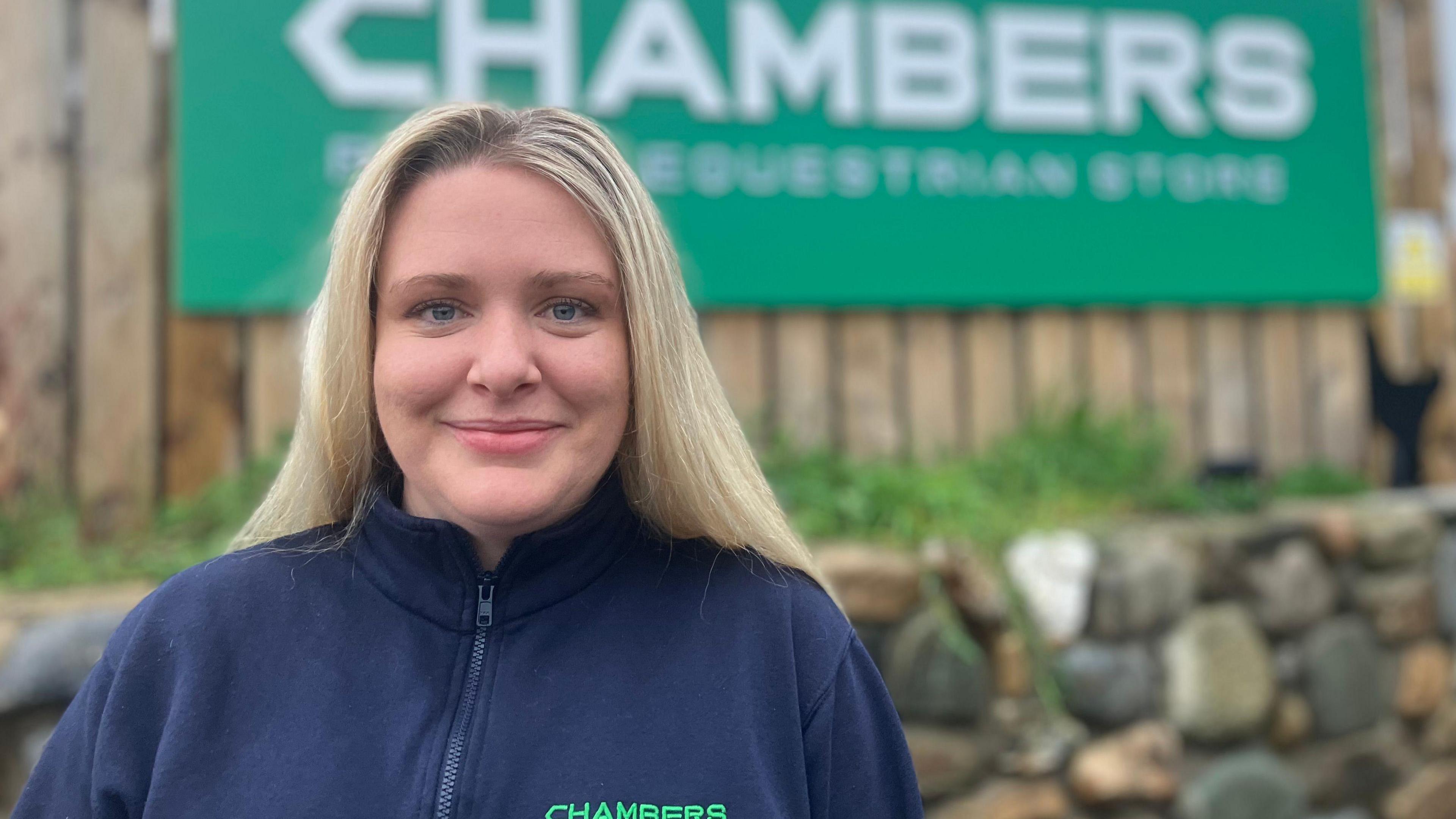 Rachel Chambers is wearing a blue fleece. She has long blonde hair and is standing near a green chambers sign.