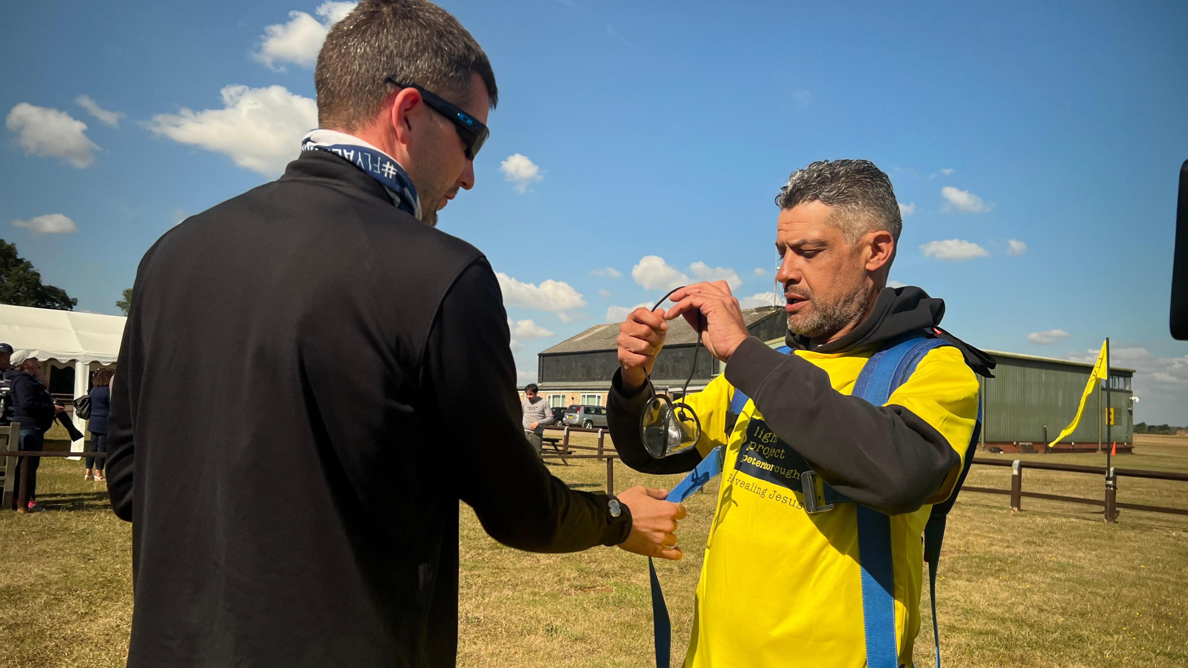 Dan Perkins wearing a yellow T-shirt and a blue parachute, and standing next to an instructor in a black top