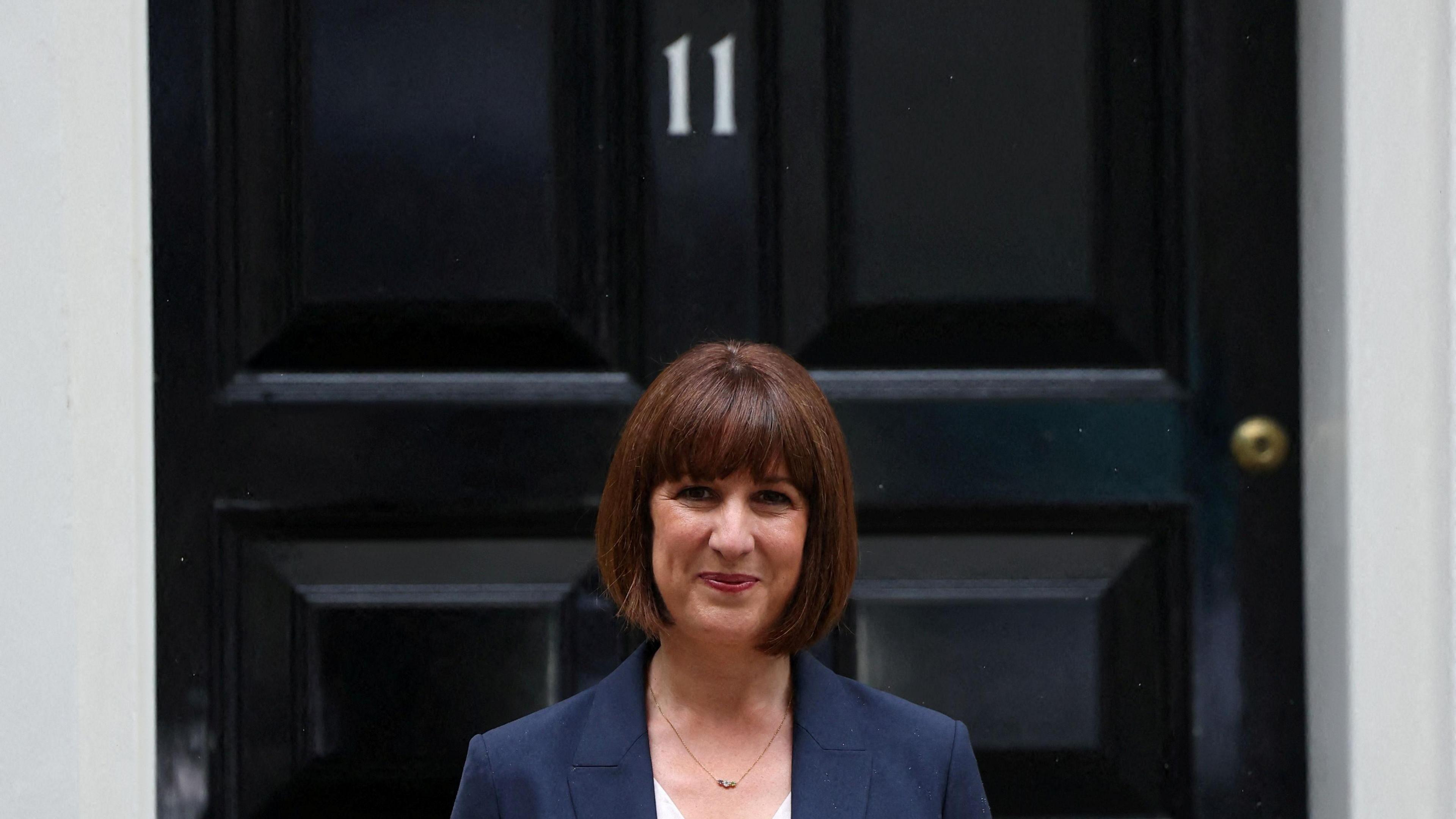 Rachel Reeves is smiling outside the door of Number 11, which is black with white number 11 on the door above her head. She is wearing red lipstick and she has straight short brown hair and a fringe. She has a small necklace on and is wearing a blue navy blazer. 