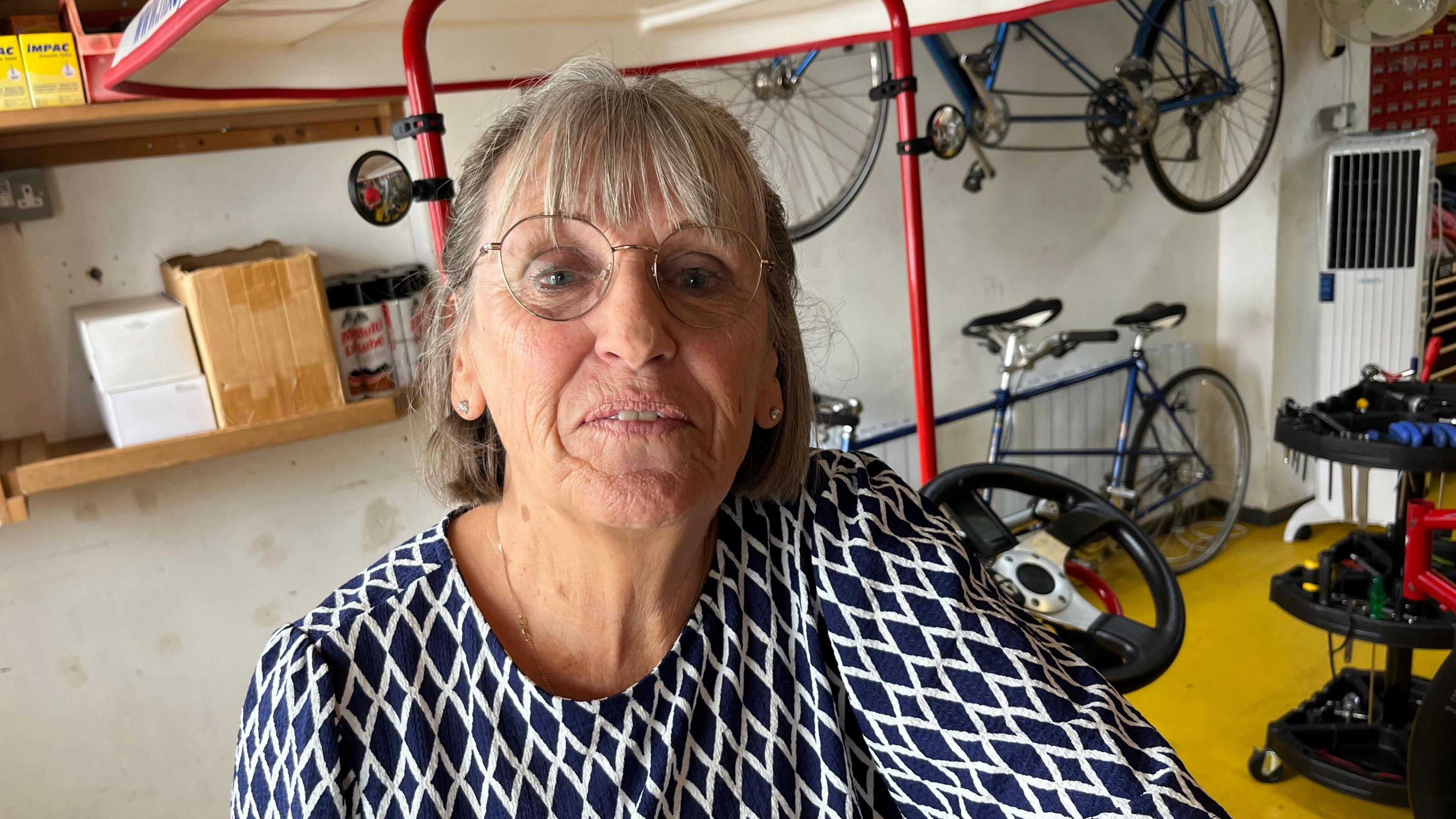 Linda Hindmarch, 74, has grey hair down to her shoulder tops, she has a fringe down to her eye brows, she is wearing a dark blue and white diamond patterned top, with a thin gold chain around her neck, she has thin gold framed glasses on too.