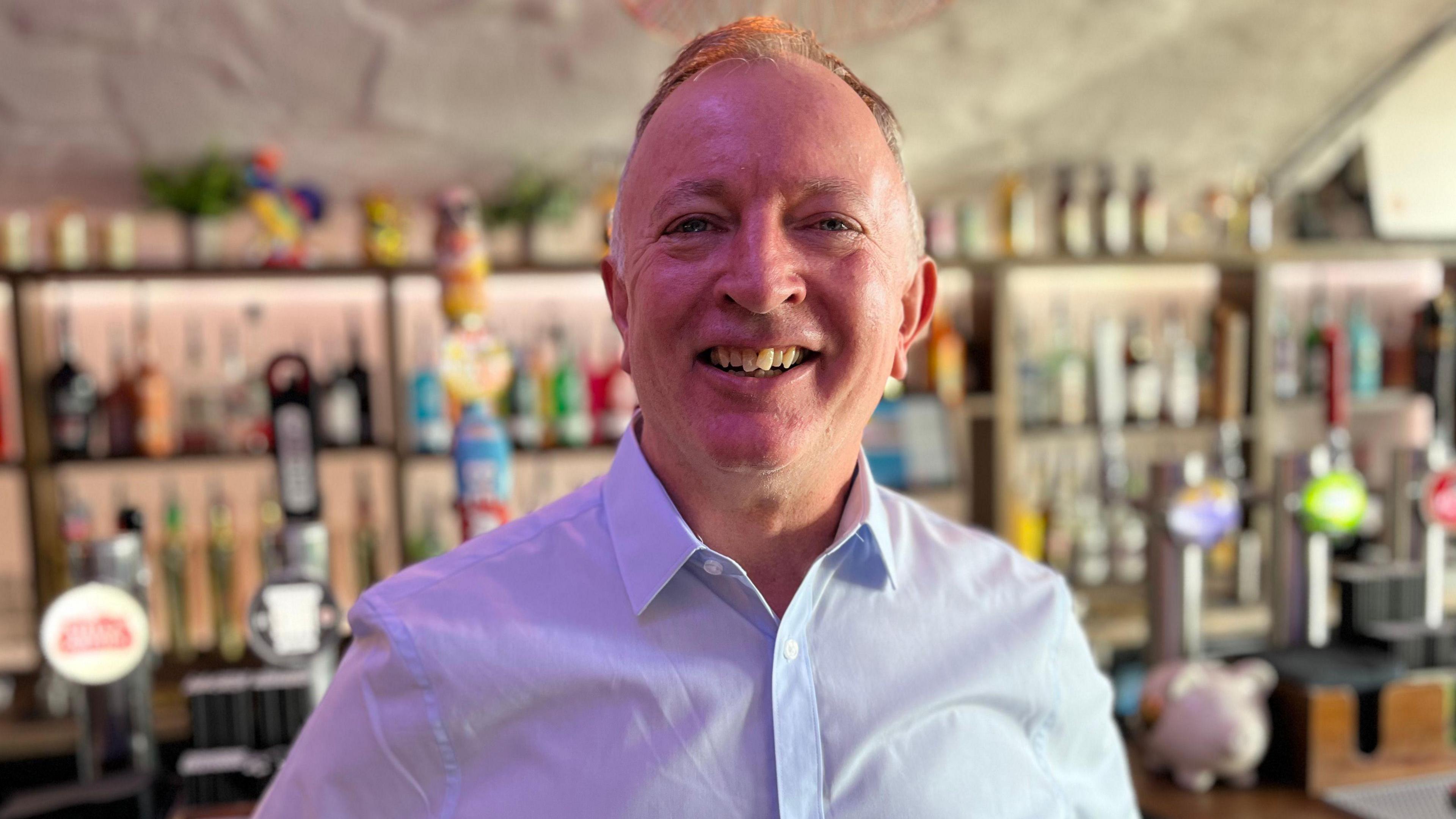 A man with a blue shirt on smiling at the camera in front of a bar. 