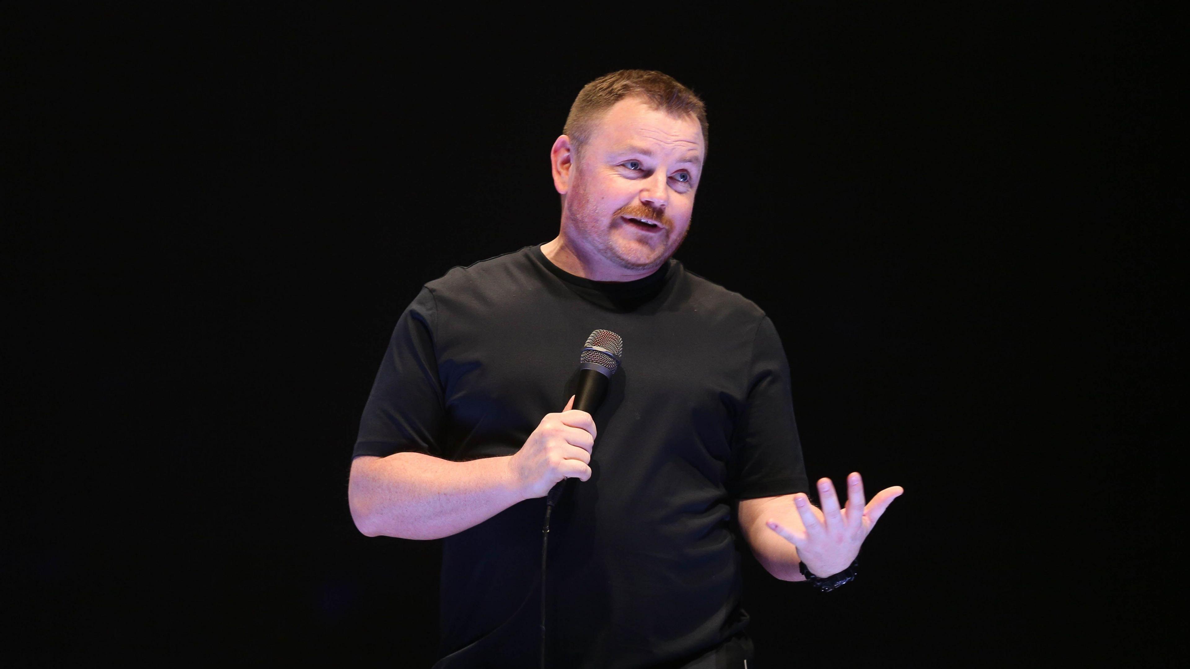 John Meagher stands, wearing a black t-shirt, alone on stage, holding a microphone 