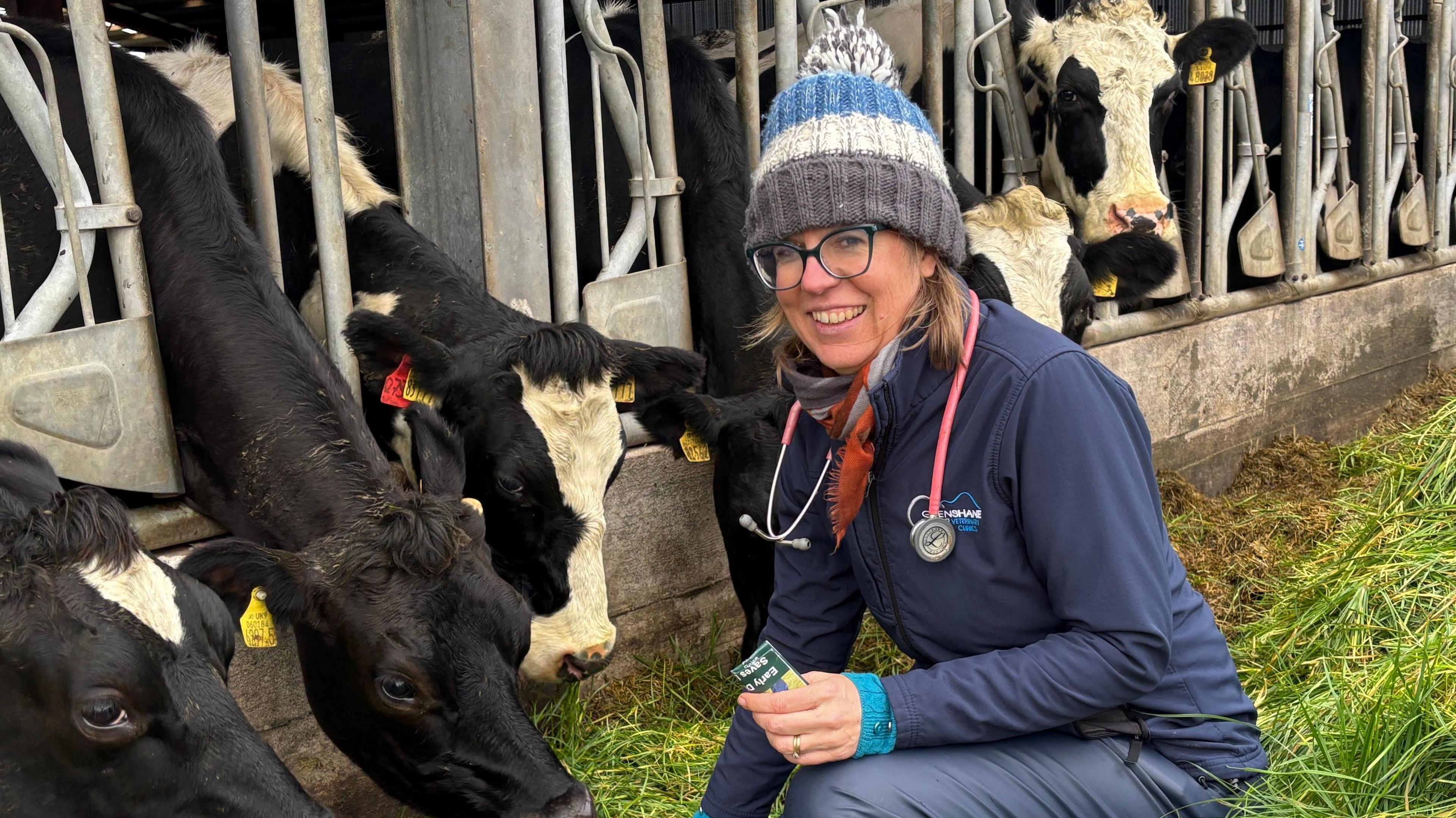 A vet crouches alongside some cows who are feeding on grass. She is dressed in a navy top and waterproof trousers with a colorful striped bobble hat. She has a stethoscope around her neck and a wide smile.