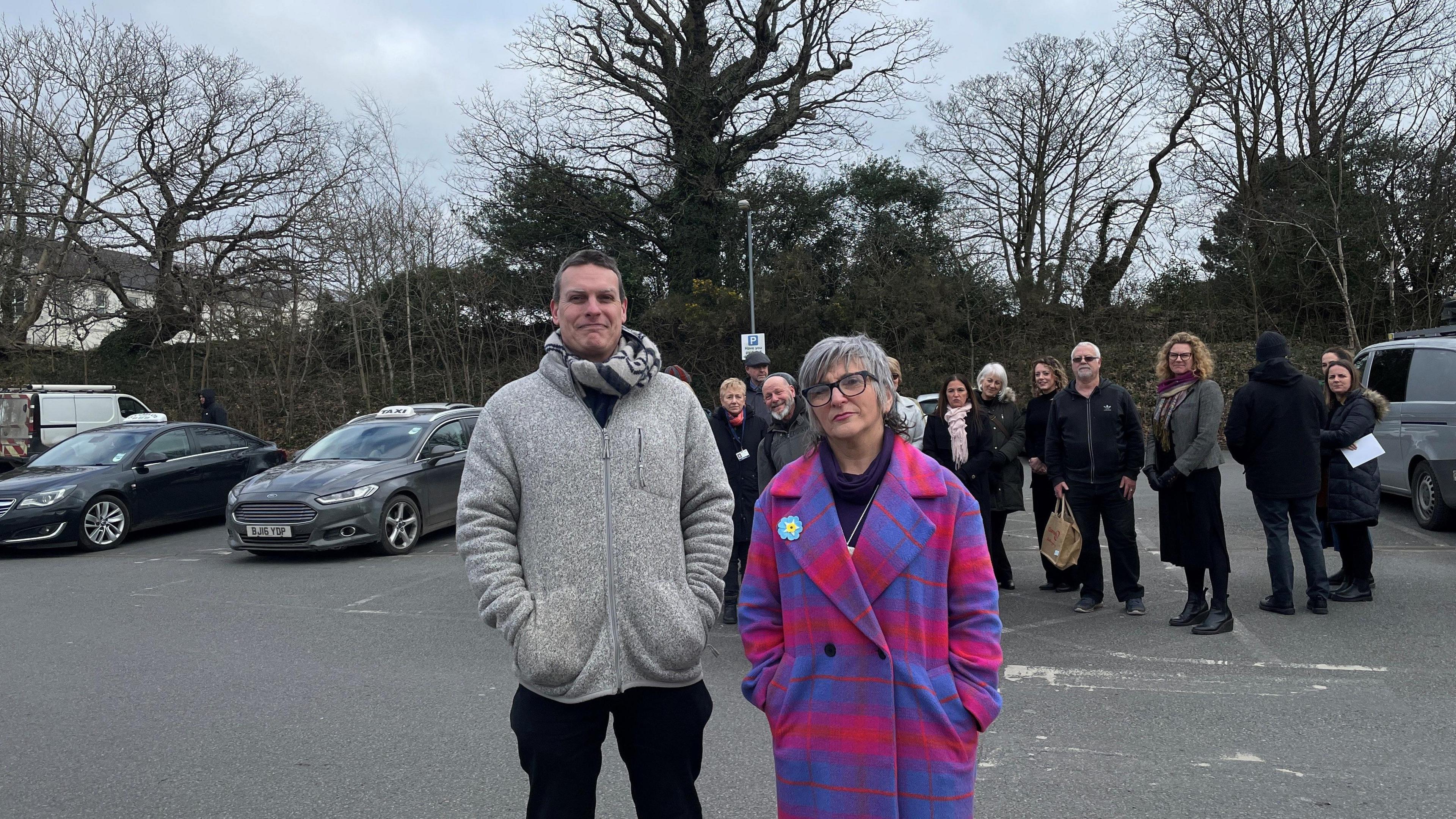 Two shop owners stood in a car park. They are looking straight at the camera. One man is in a grey jumper. The lady is in a purple and pink coat. There are multiple people behind in the background along with some cars.