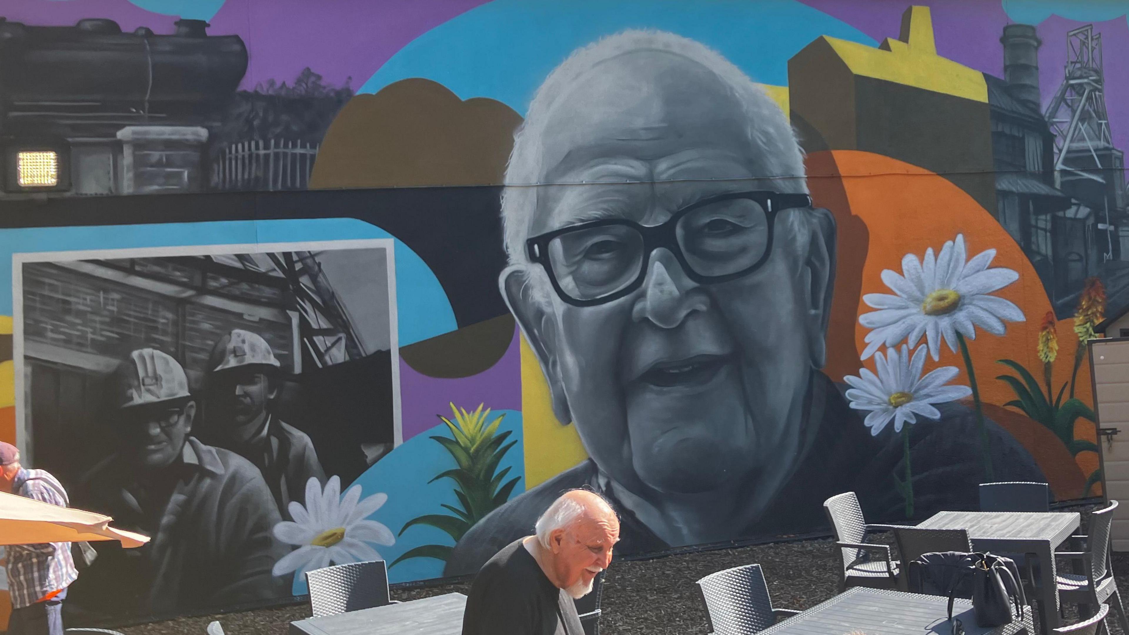 A mural featuring the greyscale image of a man's face surrounded by colourful representations of flowers and local landmarks