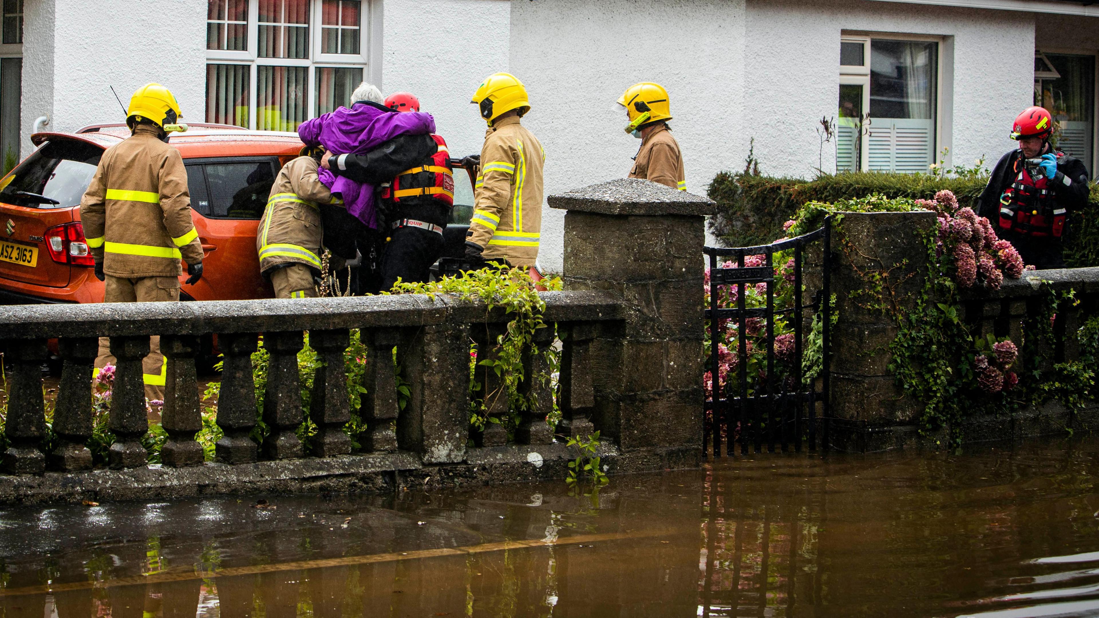 An elderly person is being lifted by fire service into a car as water flows around her home. 