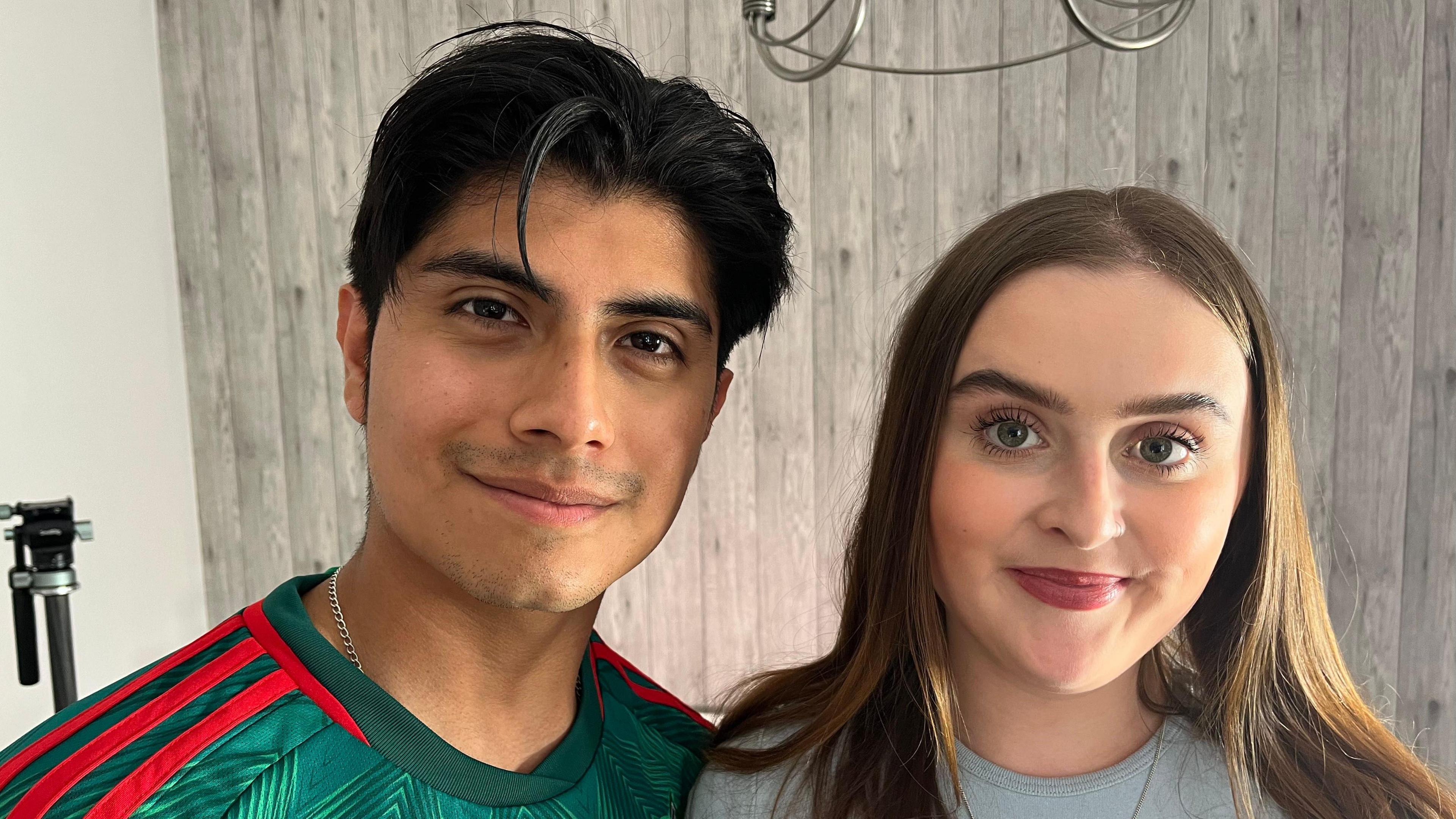 Diego - with black hair and wearing a green football shirt with three red stripes on the shoulder - and Holly - with long brown hair and wearing a grey top - both stand smiling.
