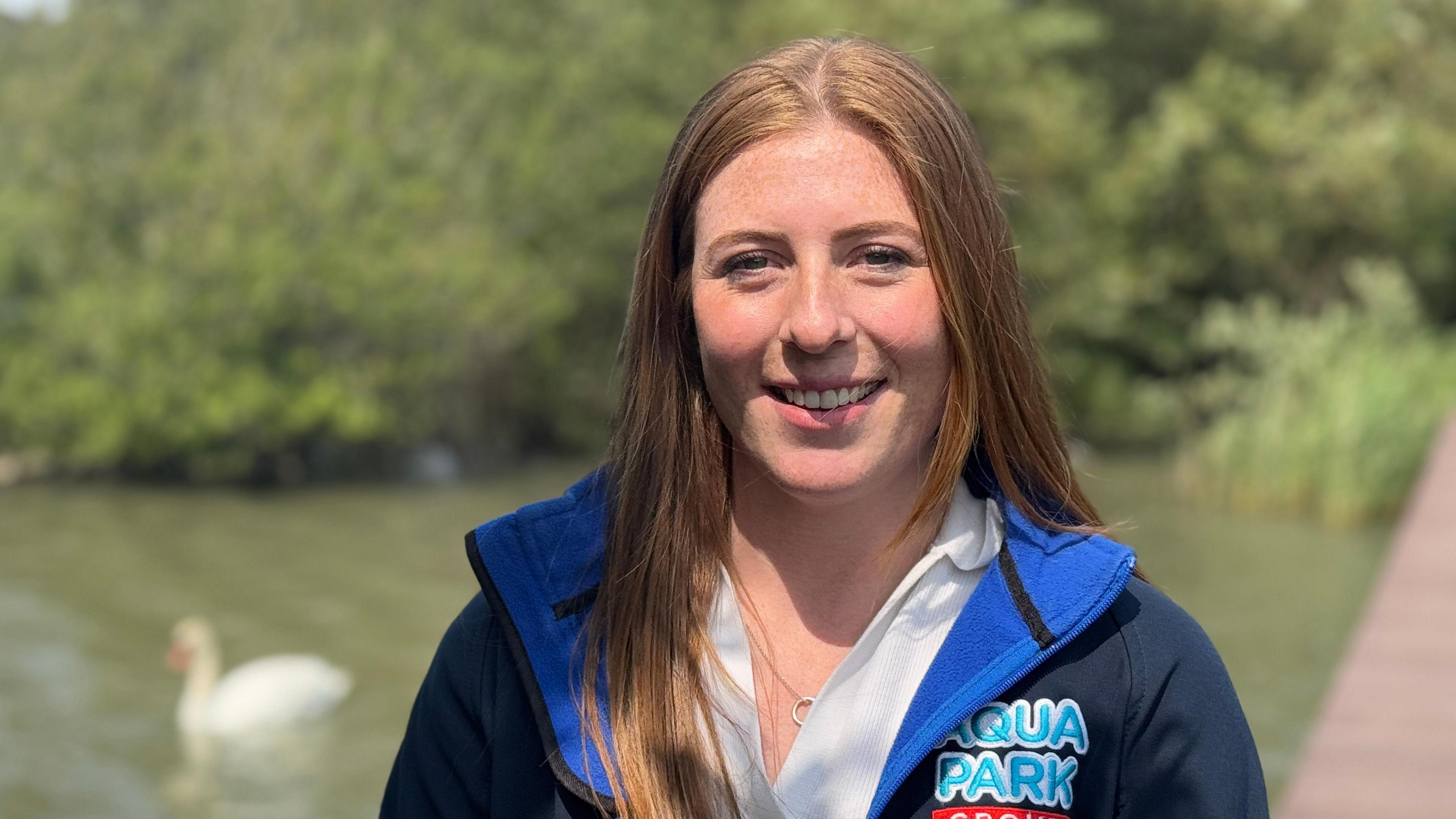 Lucy Mott wearing a blue fleece jacket. She is smiling at the camera. Behind her are trees as she stands in a country park. She has long gingery/blonde hair which glistens in the sunlight. Behind her is the lake with a swan on it - it is slightly out of focus, as she is in focus.