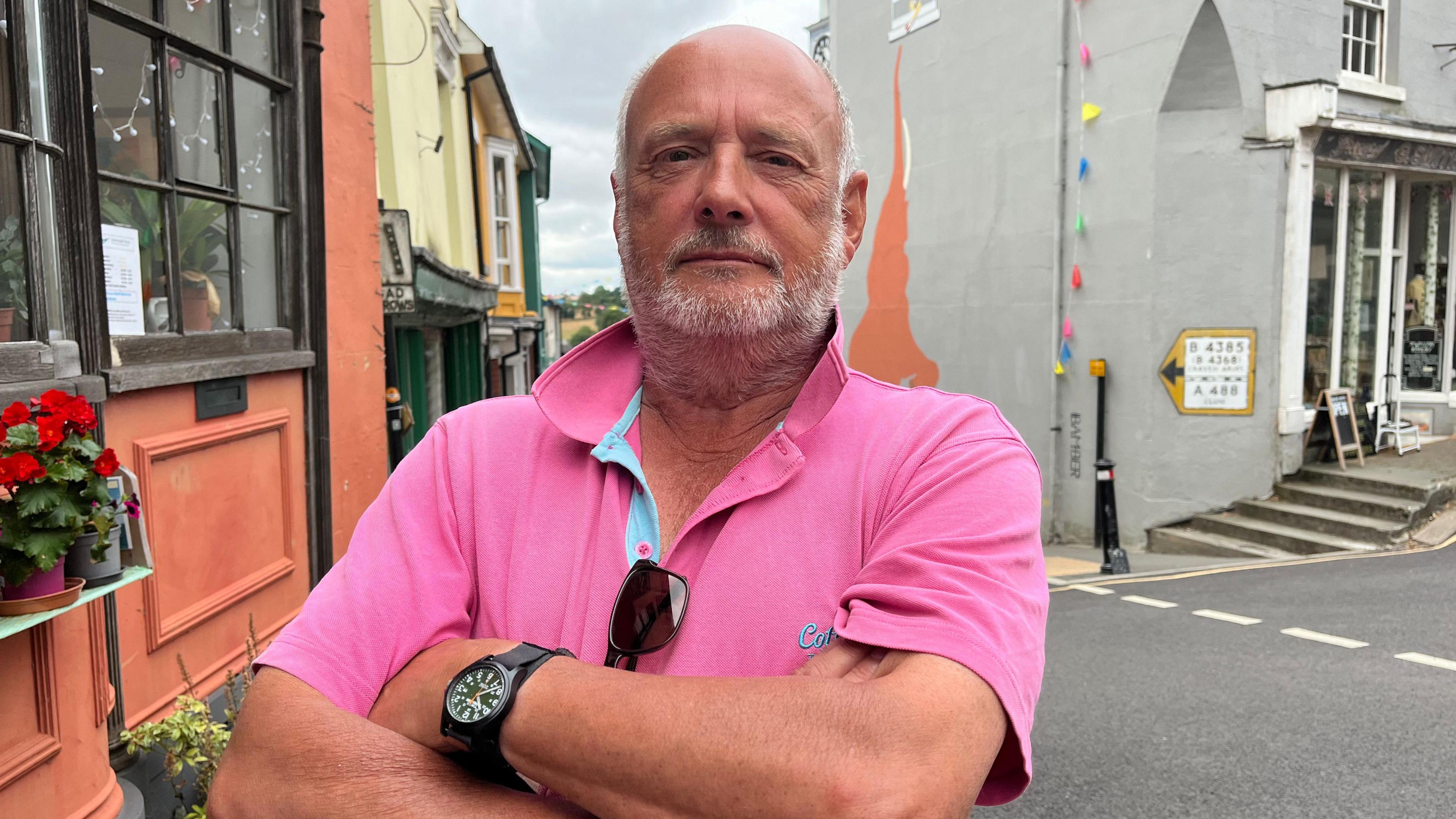 Trevor pictured looking into the camera with folded arms and a serious expression. He's wearing a bright pink polo shirt and a black wristwatch. He's stood on a pavement outside a shopfront that's painted orange.