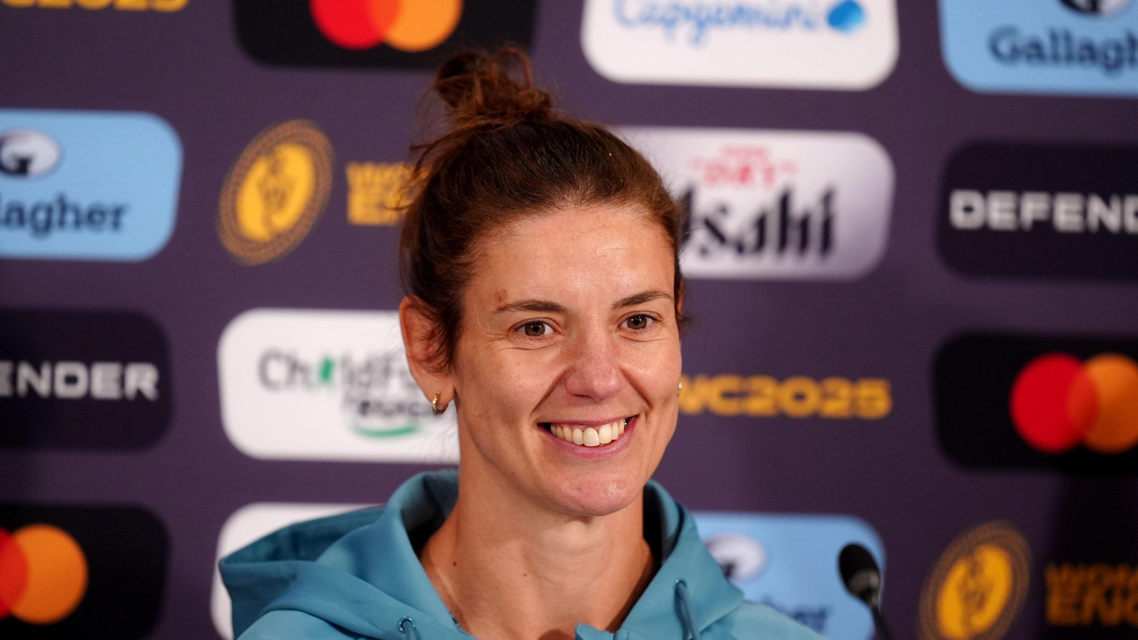 England coach Sarah Hunter at a press conference. She has long brown hair, which has been tied up and is wearing a blue hoodie. She is smiling at the camera. A small microphone stands in front of her.