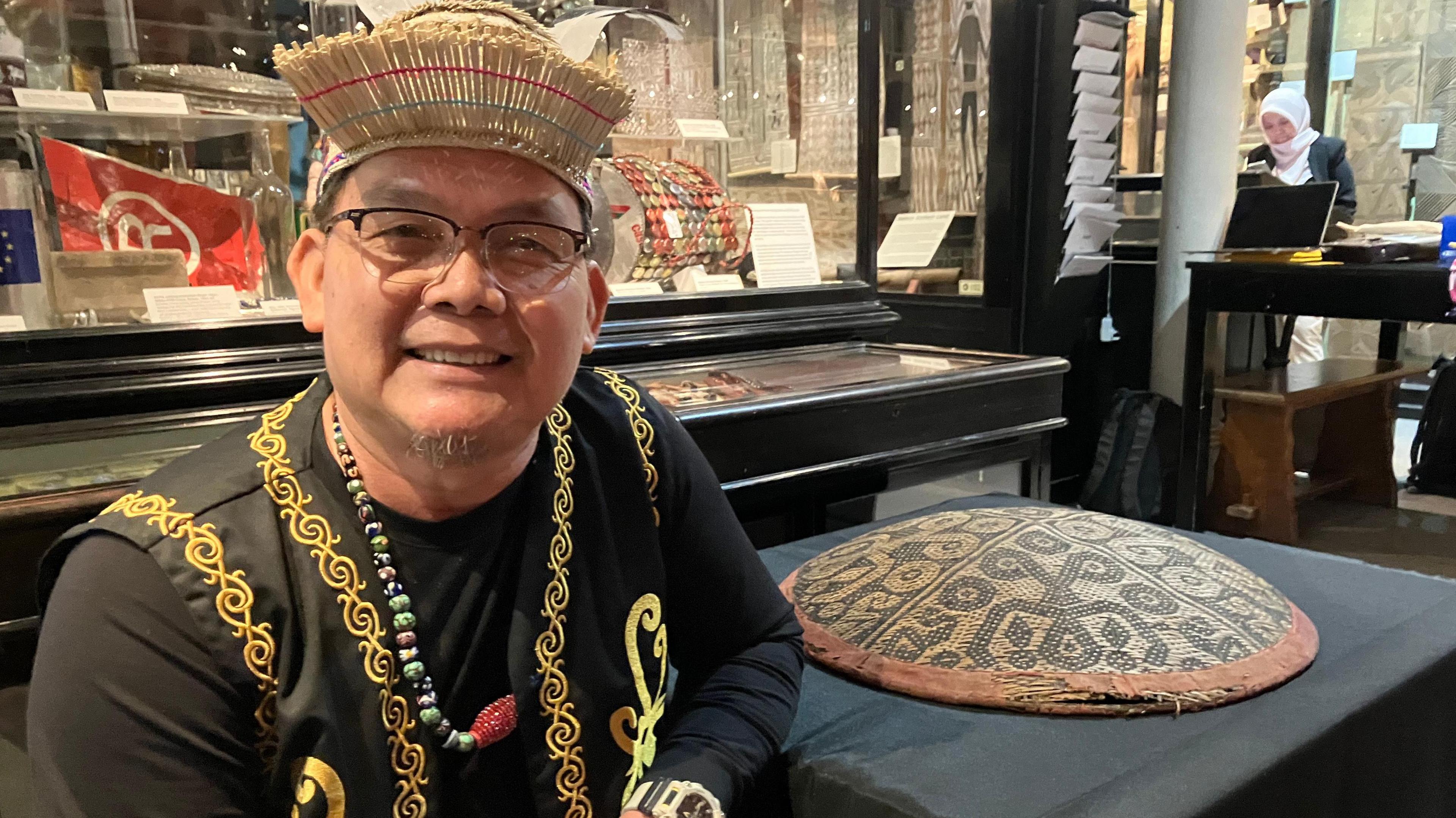 A representative of the Kenyah Badeng Association in traditional dress sits in the Pitt Rivers Museum alongside the hat, which is on a table with a black tablecloth.