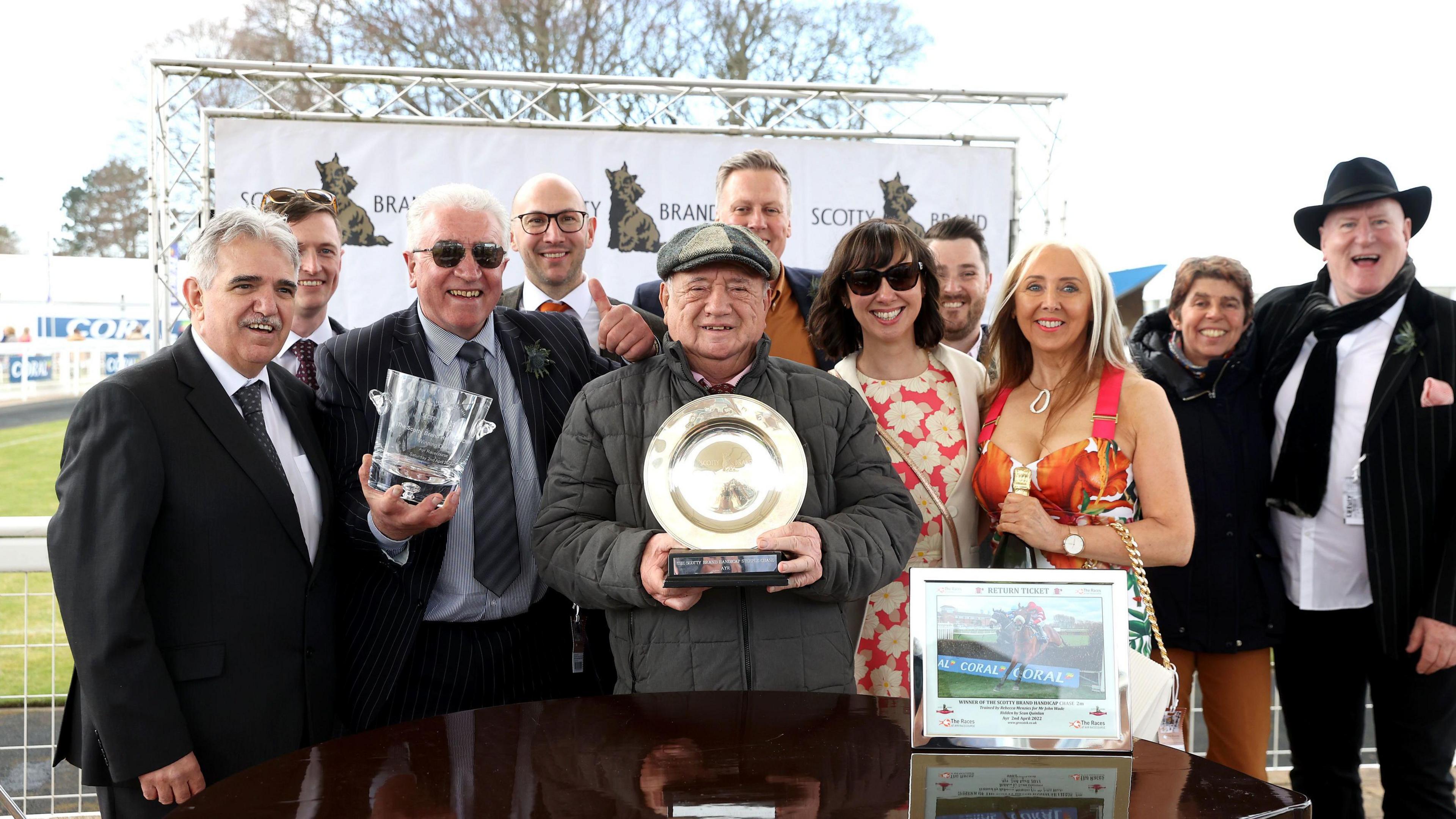 John Wade standing in the centre of a crowd at a horse race. He is holding a winner's plate. He is wearing a flat cap and coat.