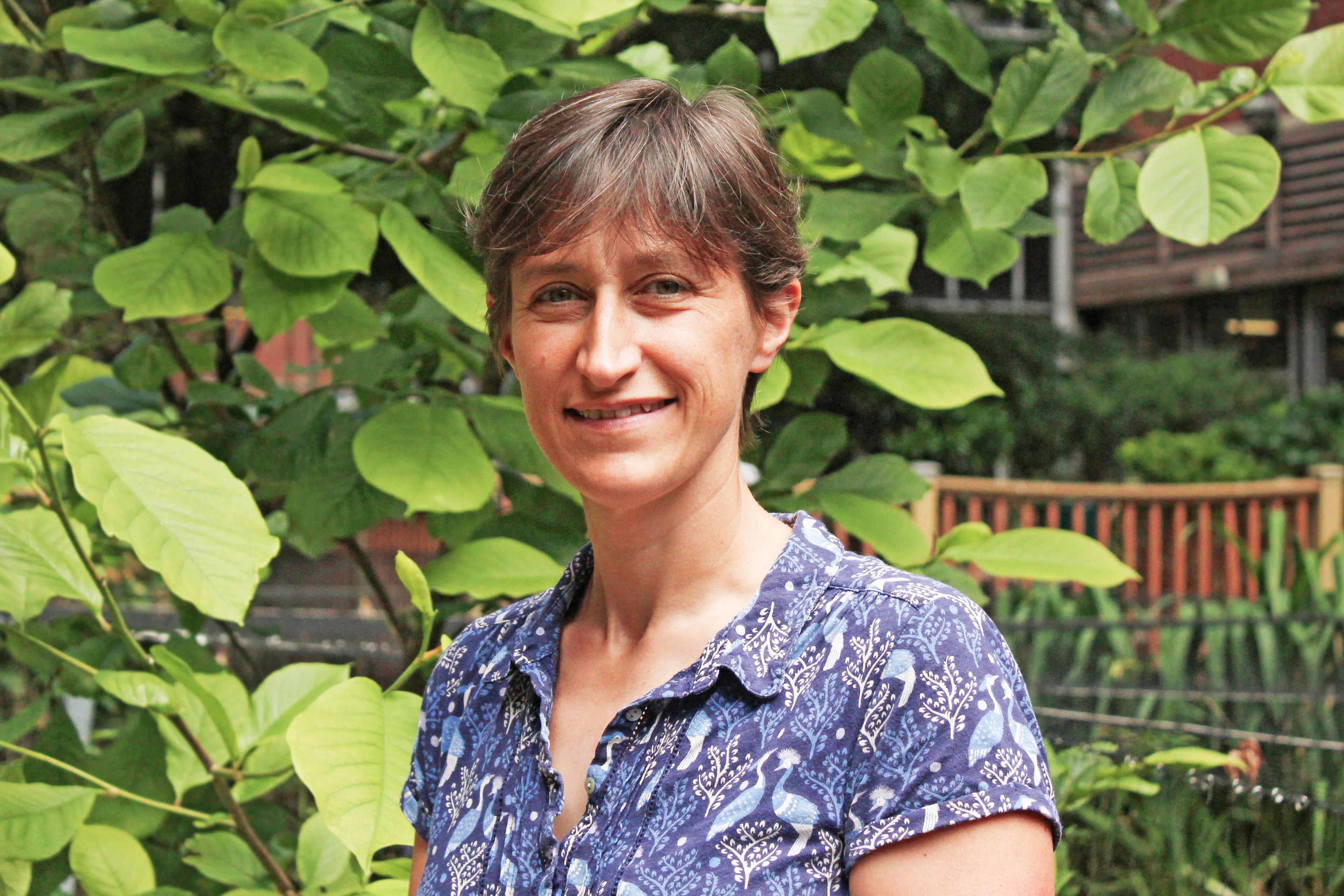 Cleo Kenington, wearing a blue patterned shirt, smiles at camera in front of green foliage