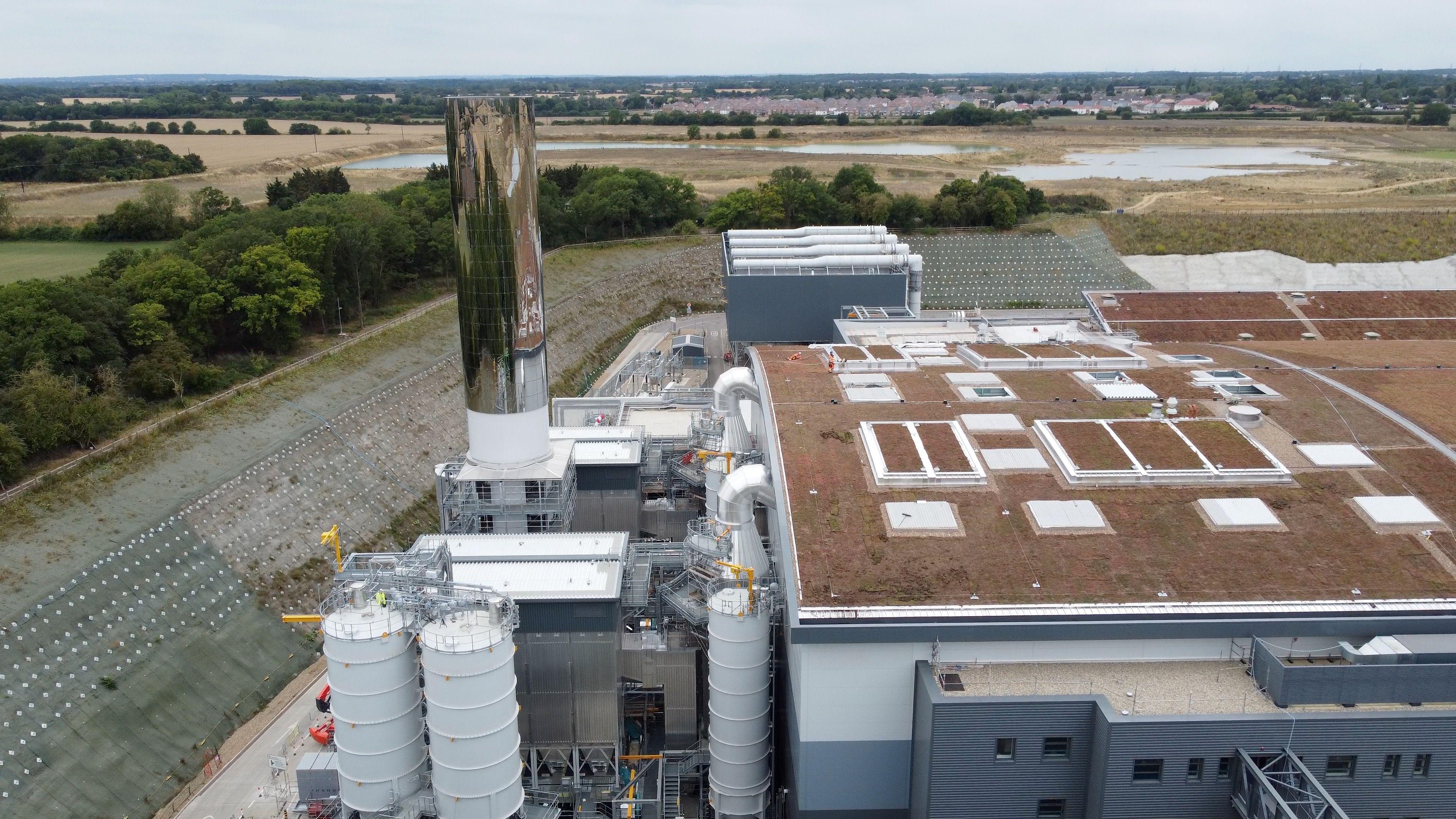 An incineration plant with a big curved building and a large chimney stack with a mirrored metal cover surrounded by green countryside with houses in the distance.