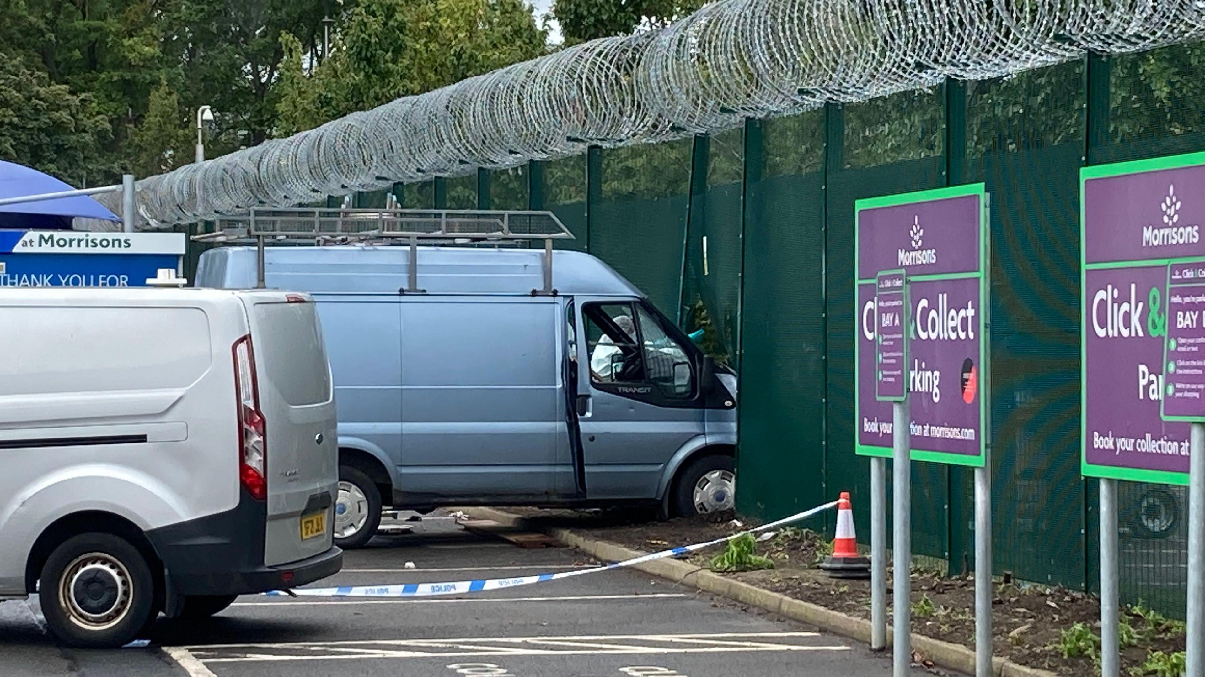 A van which has been driven into a fence in Edinburgh. The van is light grey in colour. It has a silver roof rack. The front has buckled the fence, which is green and topped with barbed wire.