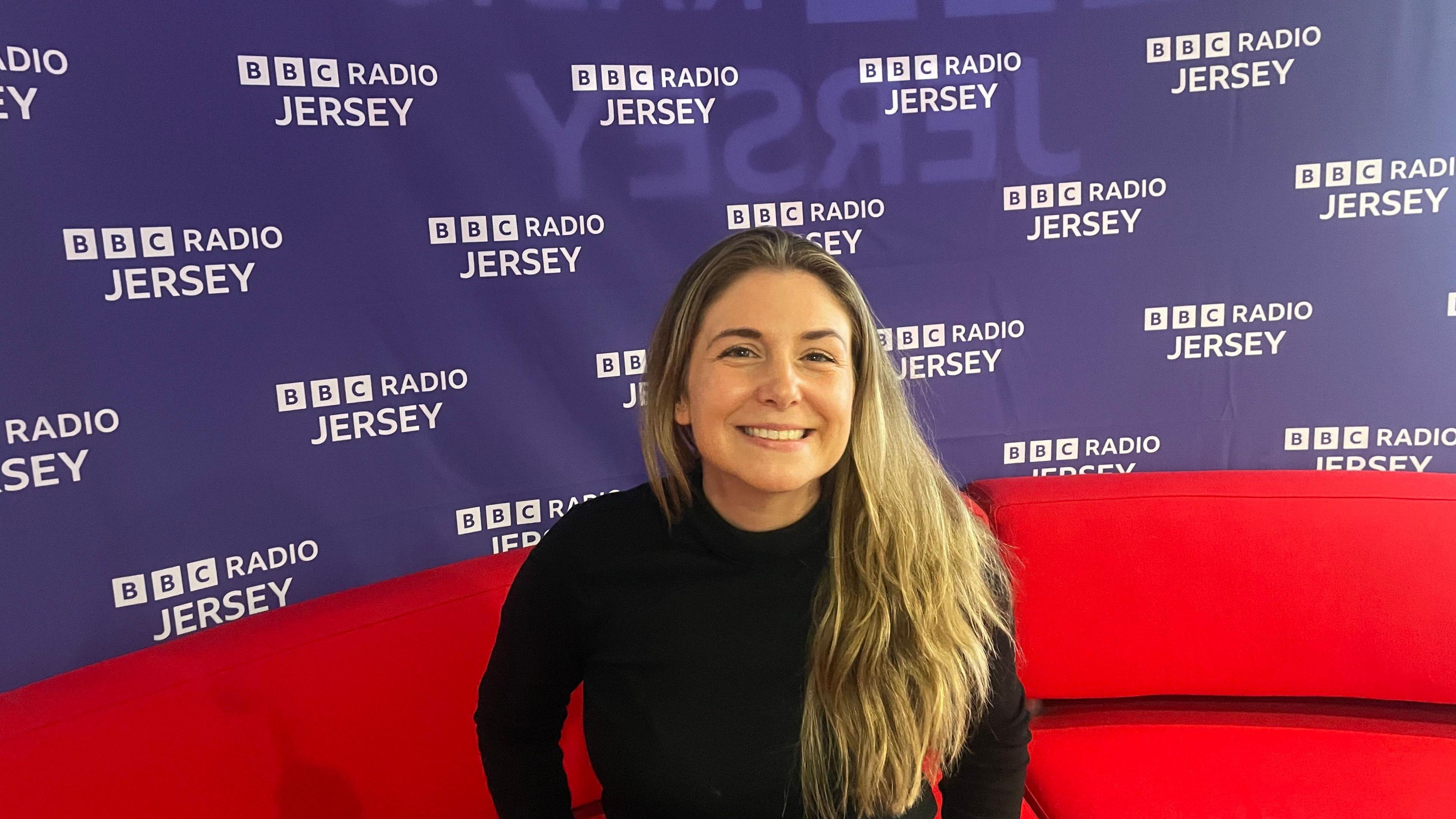 Lesley Mourant sits on a red sofa in front of a Radio Jersey branded purple wall, her long hair is over one shoulder and she smiles towards the camera.