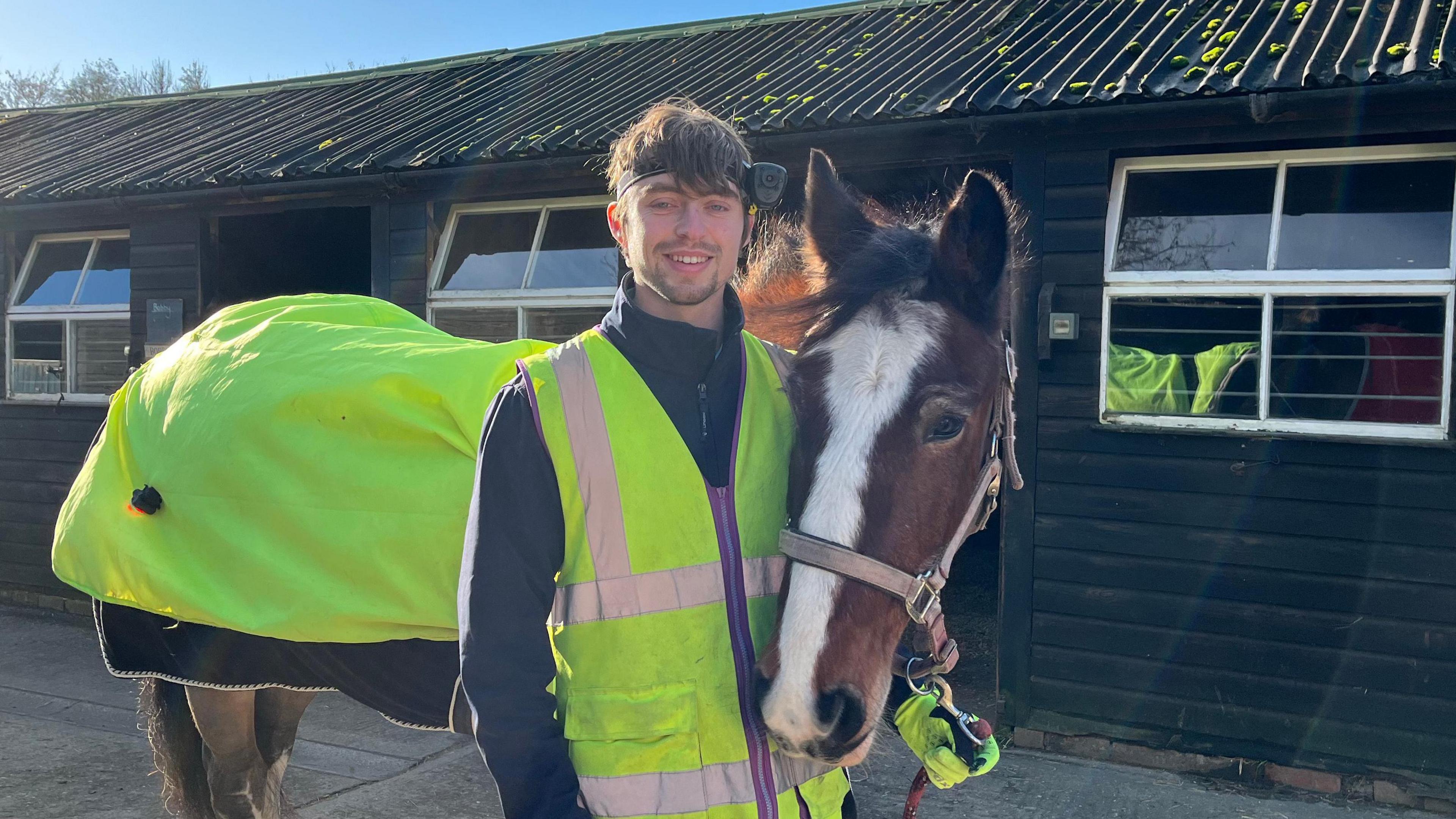 Rory Leggett holding his brown horse badger at their stable. Both Rory and Badger have high vis clothing on to make them more visible to cars