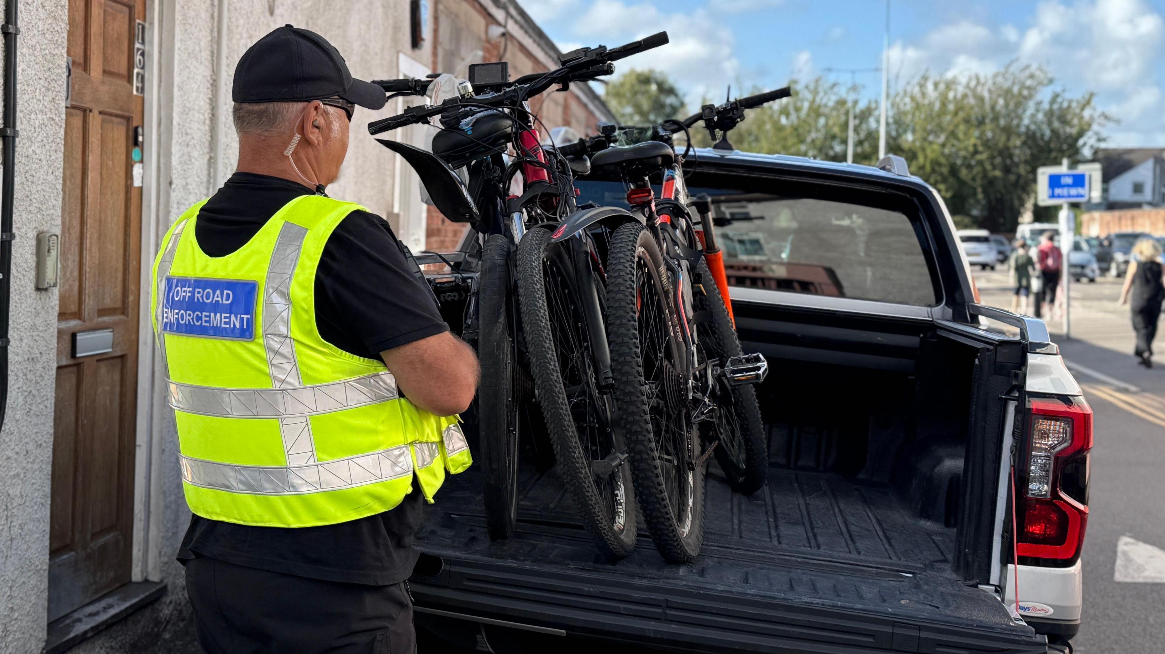 Three mountain bikes loaded into the back of a pick-up truck. Next to the vehicle a council officer stands with his back to the camera, wearing a black T-shirt and yellow hi-vis jacket which has a blue label on the back which says "Off Road Enforcement"