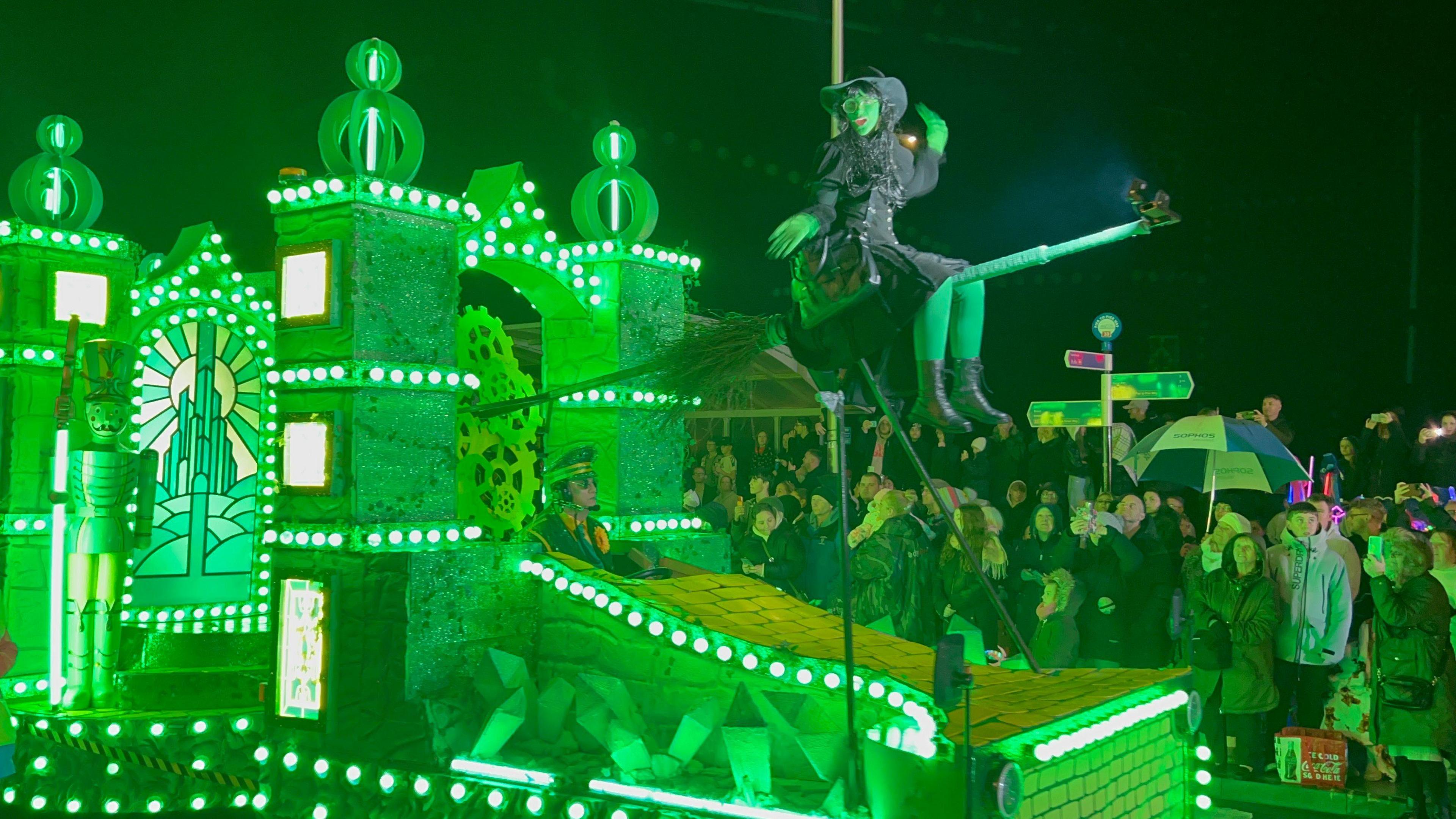 A green illuminated carnival cart with a Wicked theme. A performer wearing green body paint and a witch costume is sat on a broomstick at the front of the cart. A crowd can be seen watching.