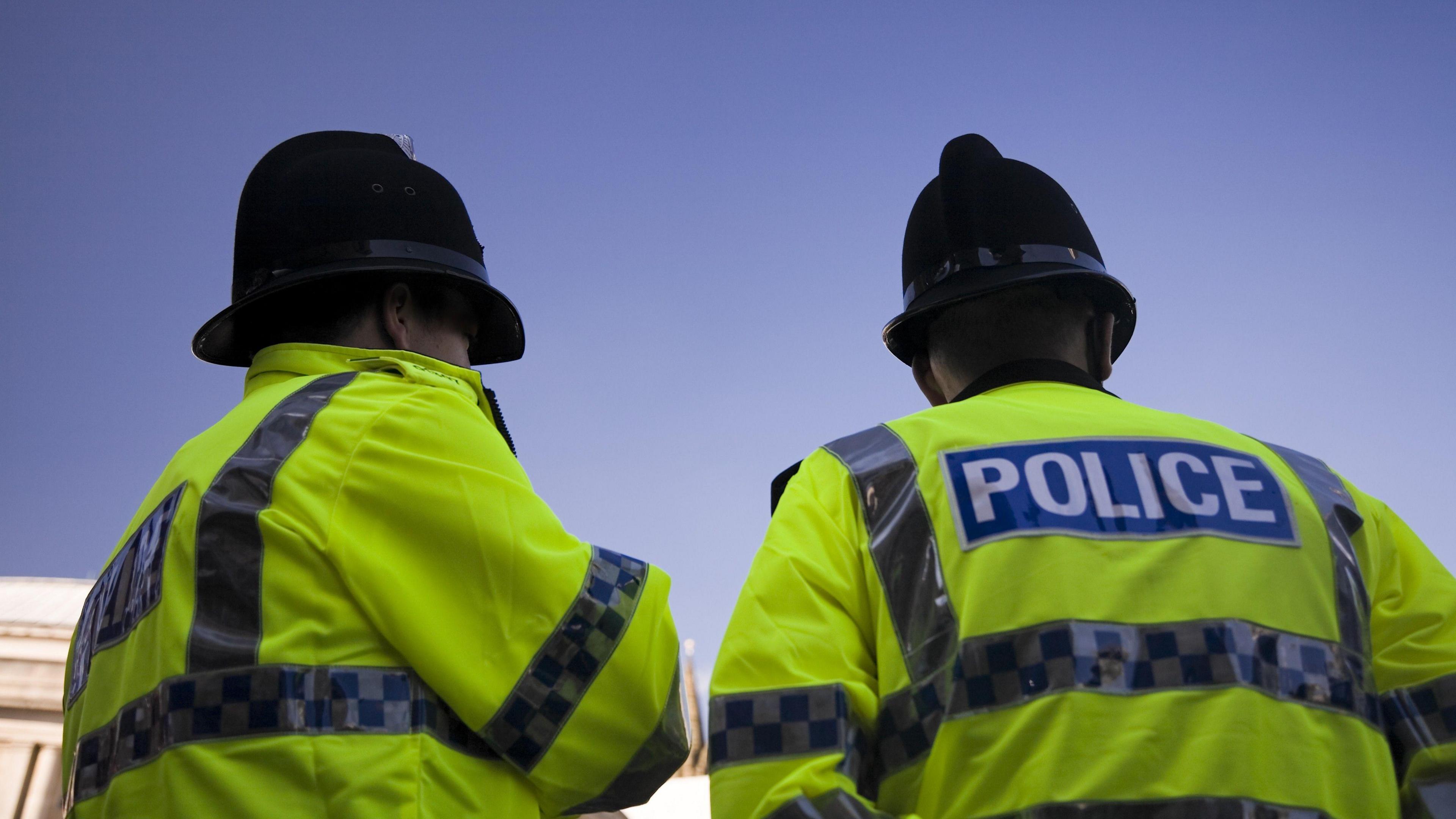 Image of two police officers in florescent jackets. POLICE is seen on the back and they're wearing black police hats. The sky is blue above them. 