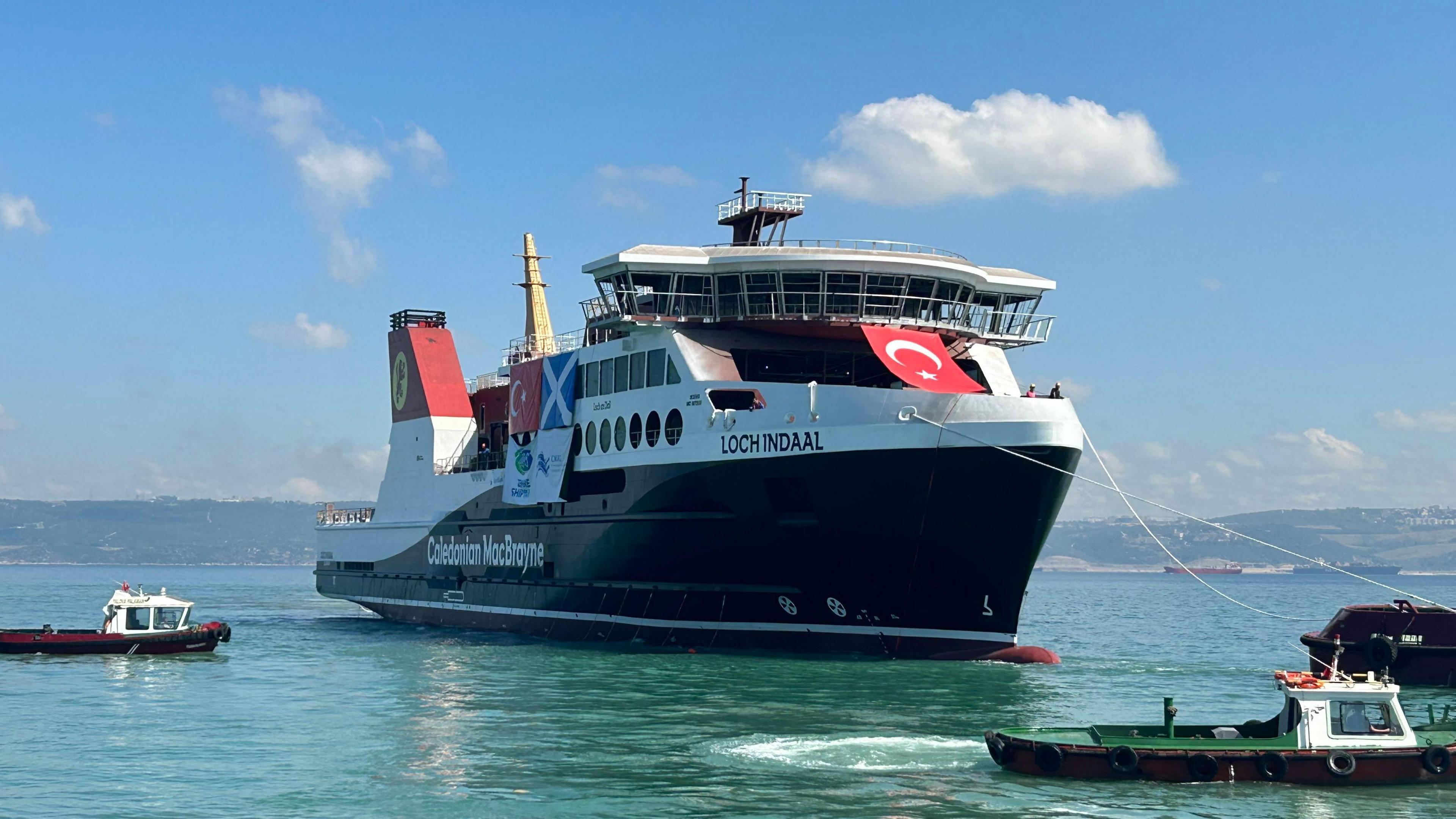 A large ferry, Loch Indaal, floating in a green sea, with tied to three smaller boats. The ferry has two turkish flags, red with white crescent moon and star, displayed on it and a saltire.