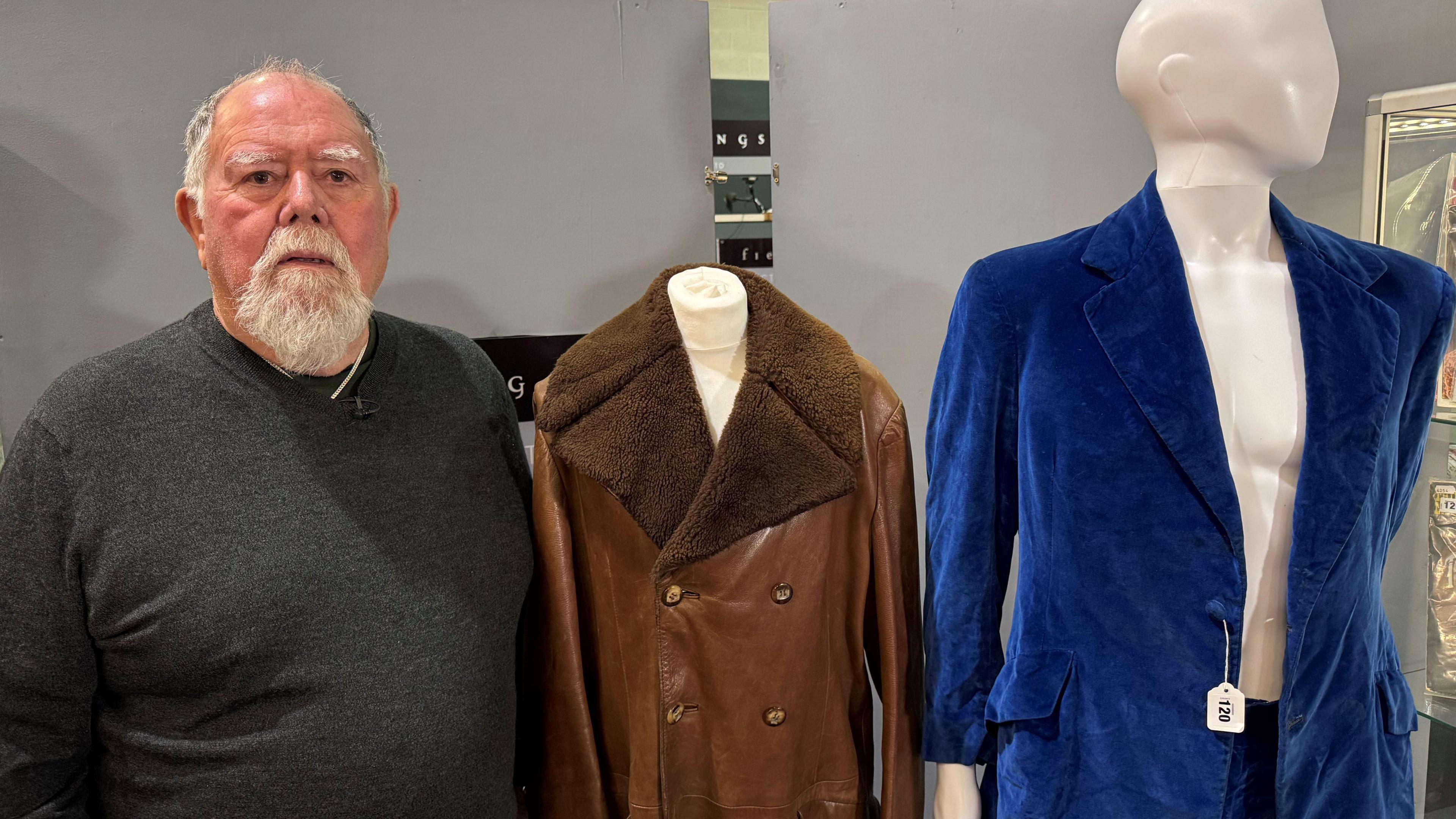 A man in a grey jumper standing next to two items of clothing. He has a grey beard and grey hair. Next to him is a brown leather jacket and a blue velvet suit which are on mannequins. 