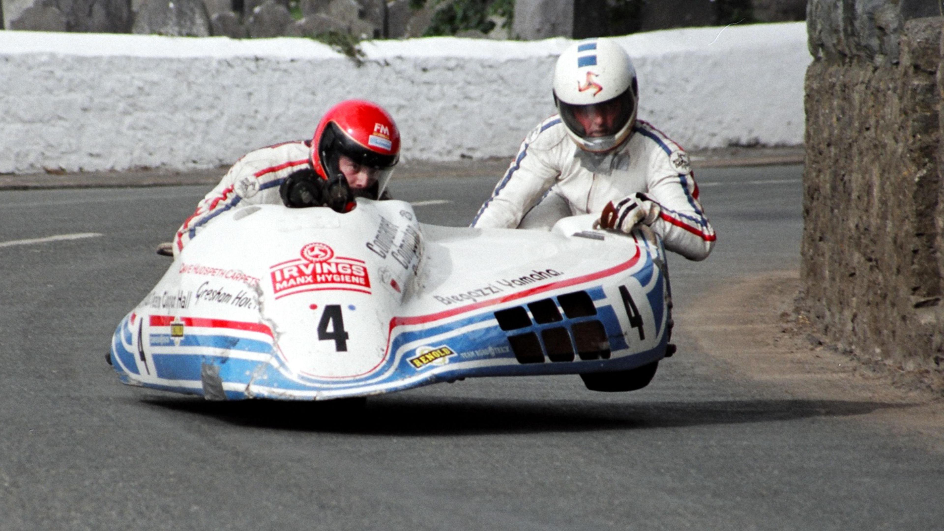 Two men in white leathers racing a white and blue sidecar outfit on a road, with the driver wearing a red helmet