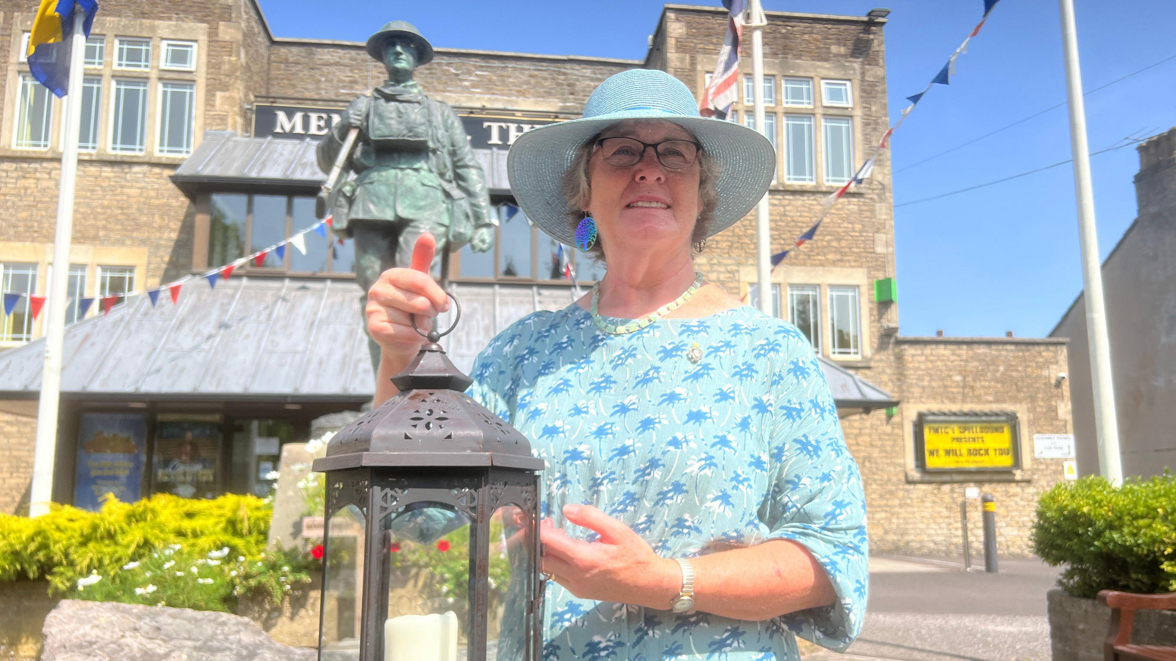 A woman in a blue sun hat and blue patterned top holds a lantern with a candle inside. Behind her is a bronze statue of a soldier. She is in an urban setting, with a large stone building and large flagpoles behind her.