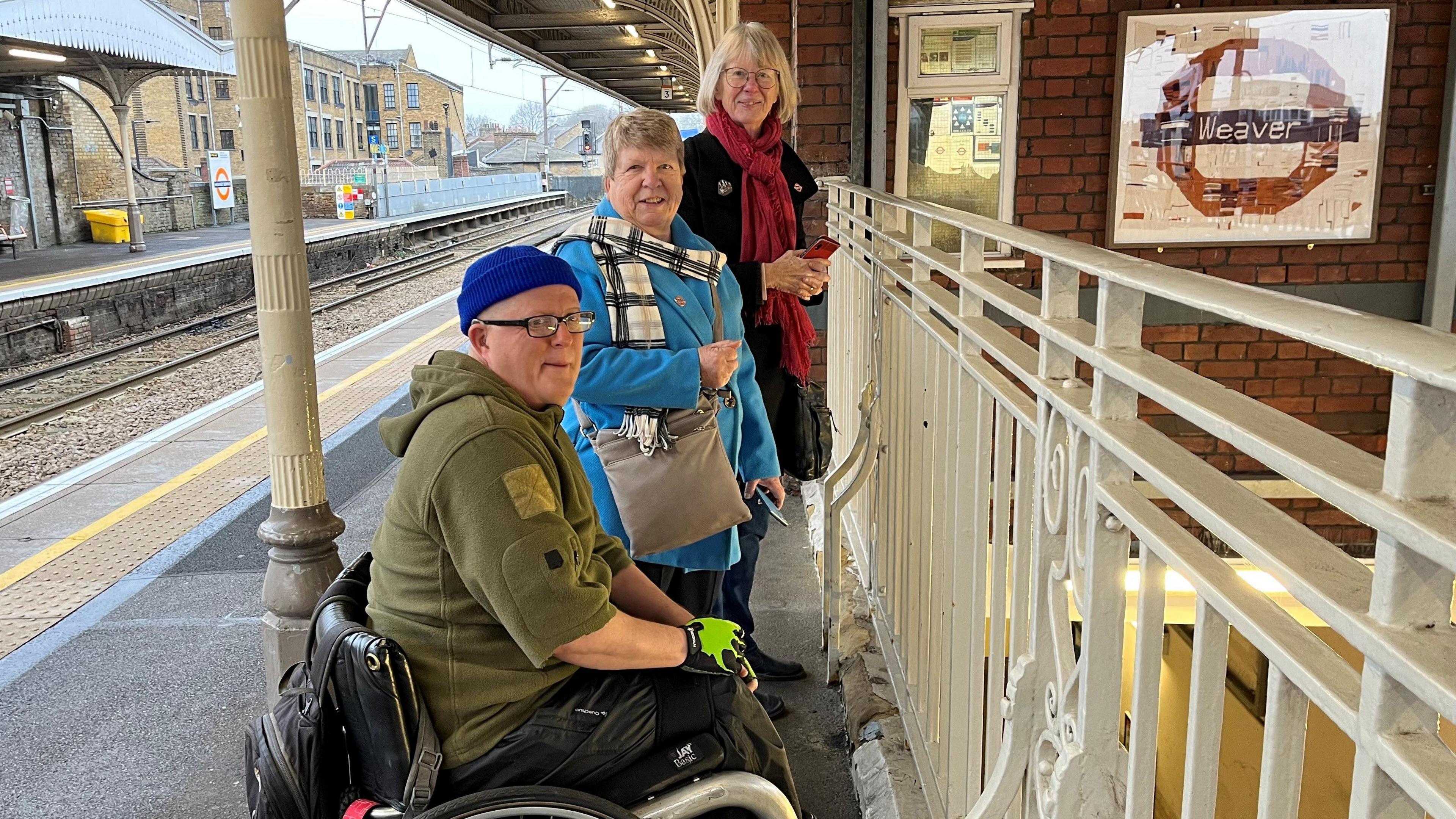 Ben, Pam and Anne are seen standing next to a railing on the train platform with the woven roundel on the wall next to them.