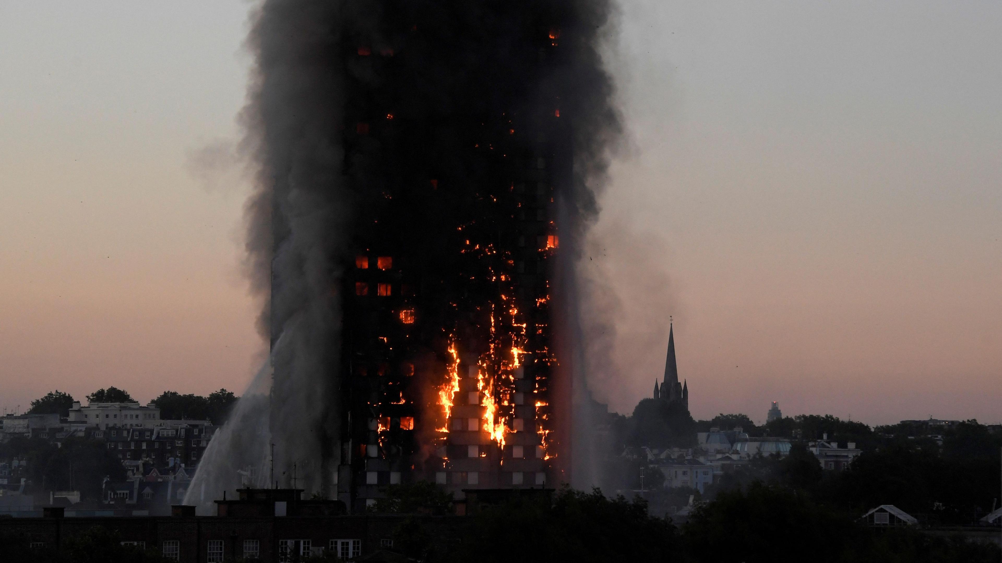 The Grenfell Tower building with fire in the windows of the bottom of the building as smoke billows above. 