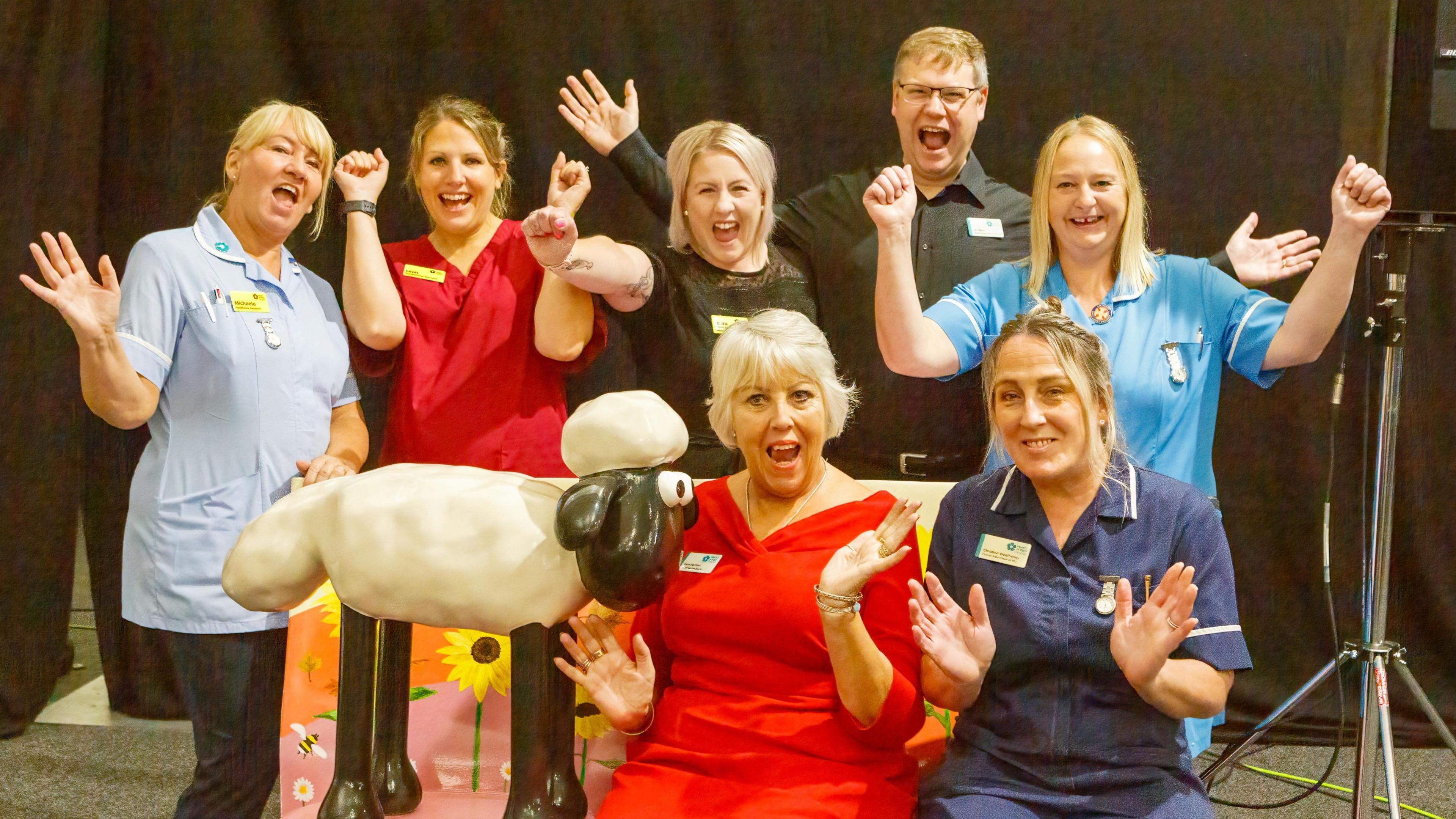 Seven hospice staff in uniform cheer as they surround and sit on a pink Shaun themed bench. 