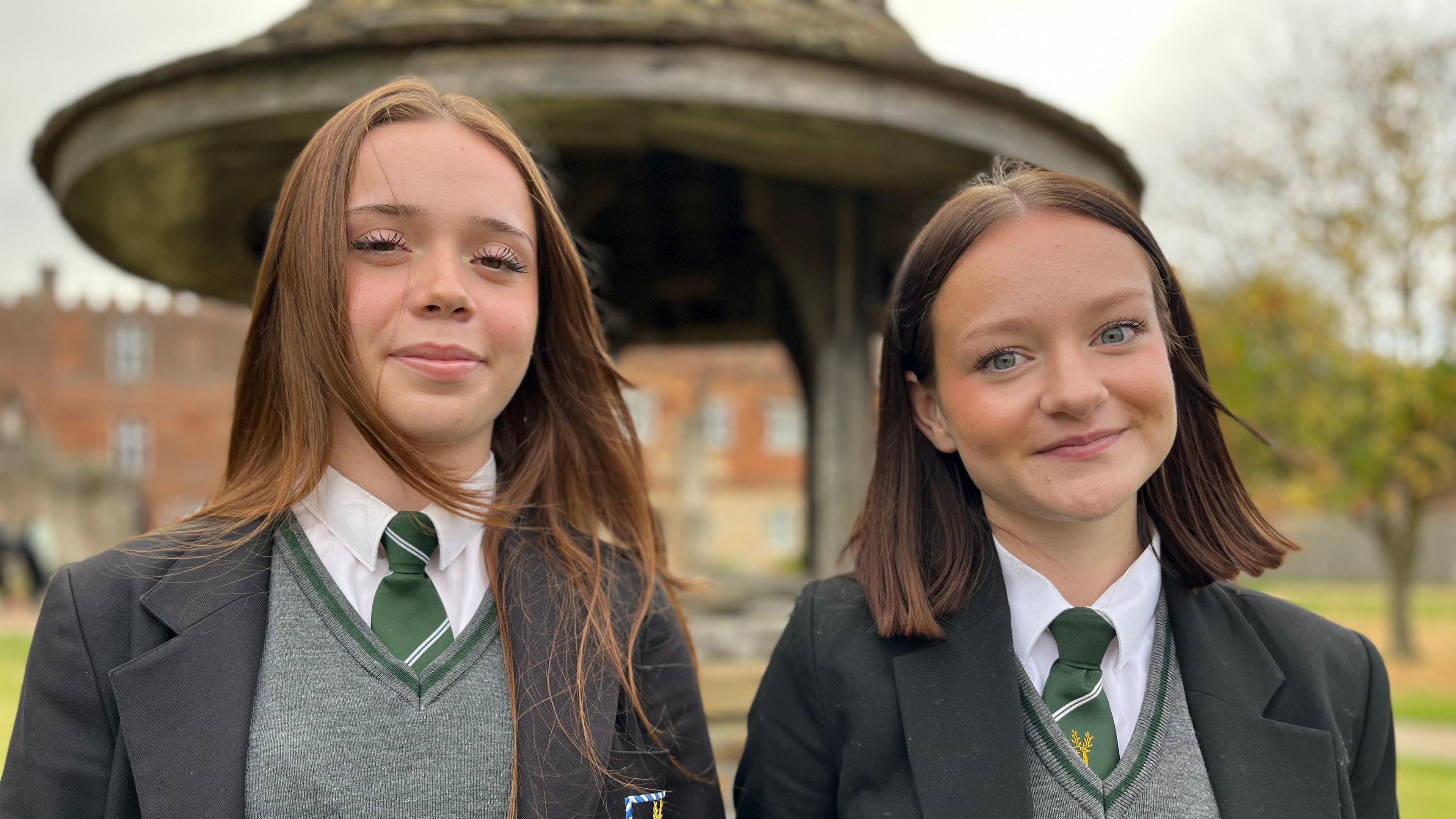 Athena and Liv are standing in school grounds and wear a black school blazer, white shirt, green school tie with a white stripe and grey jumper. Athena on the left has long red hair and fair skin, Live has shoulder length dark hair and fair skin.