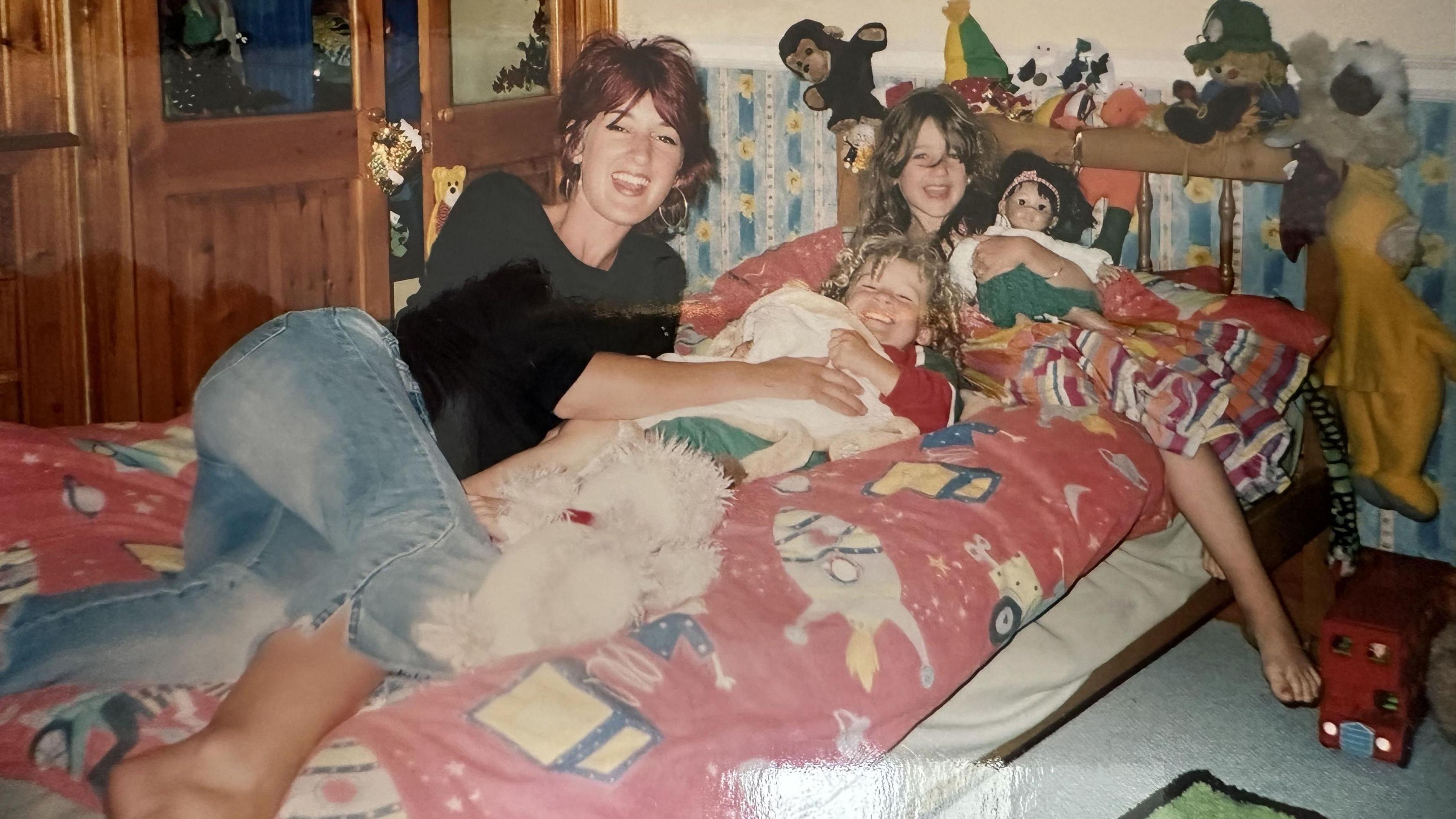 An old photo of Milo's mother, Natalie Young, sat on a child's bed in a child's room with two young children holding dolls and teddy bears. Natalie has short red dyed hair and is wearing a dark top and capris jeans.