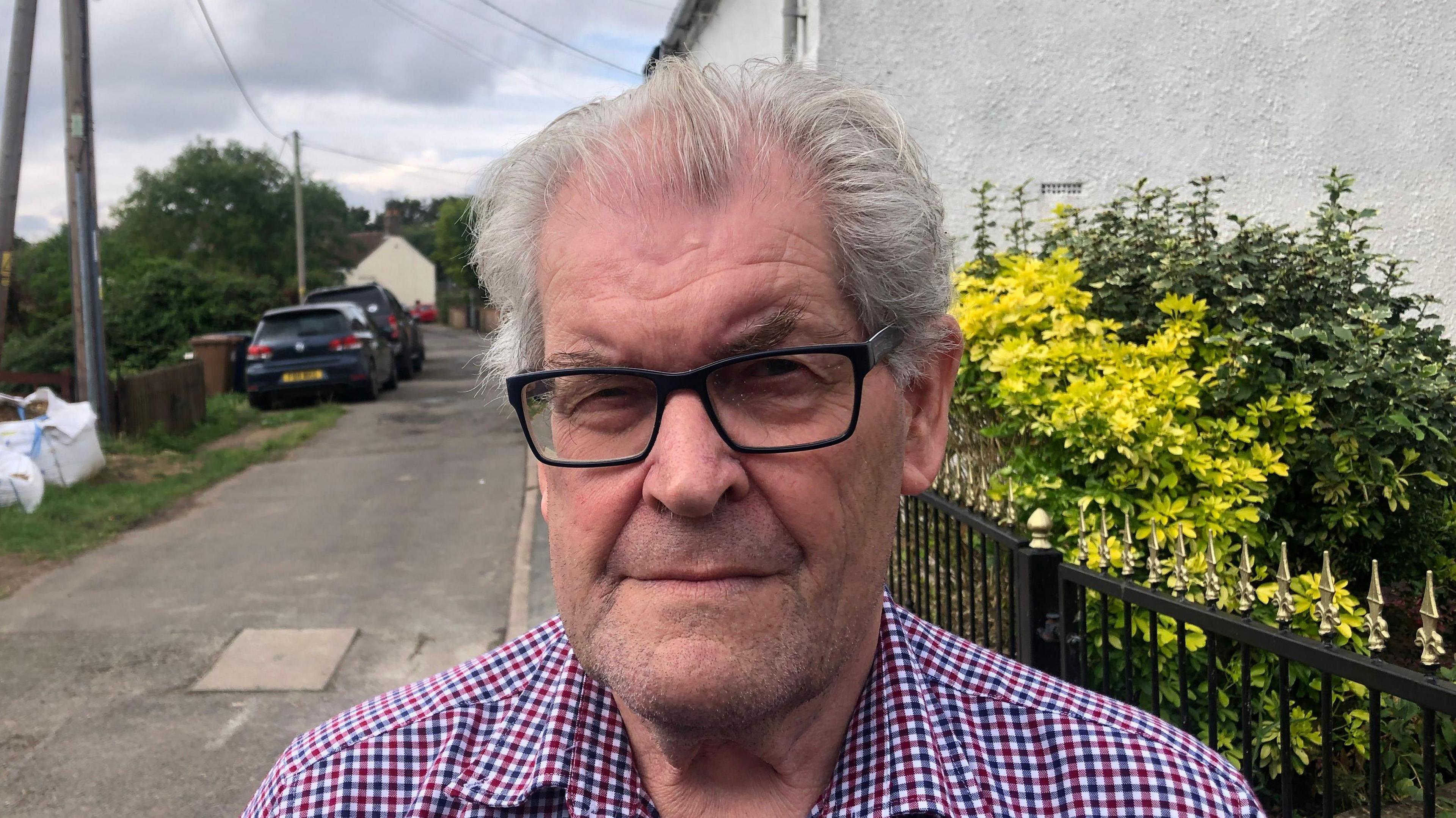 Neville Patrick looks directly at the camera wearing black glasses and a chequered shirt. He is photographed outside with residential houses behind him and a pathway with two cars parked on the right hand side. 