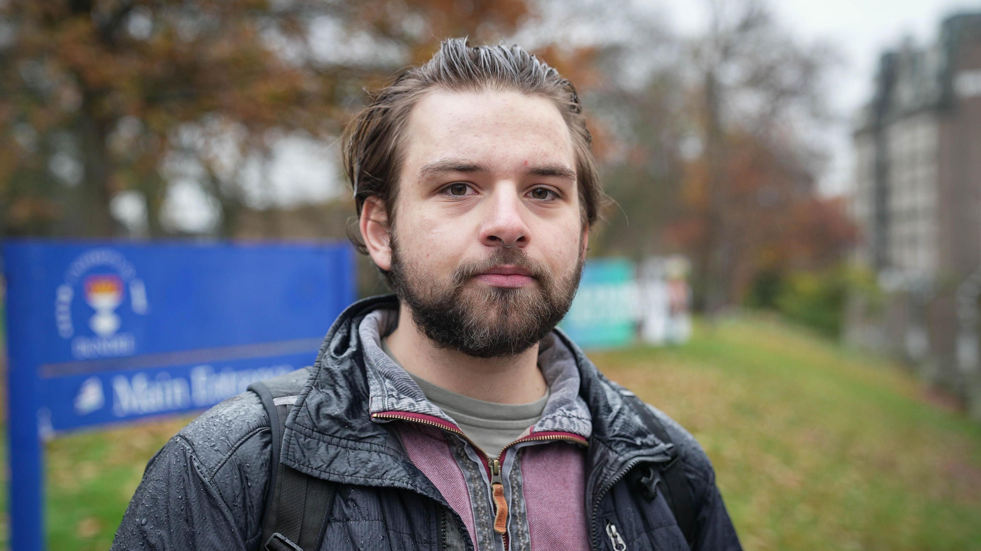 Fraser Adams has short, brown hair and a beard. He is standing outdoors on a cloudy day, wearing a rain-speckled black jacket and backpack. A blue sign and autumn trees are visible in the background.