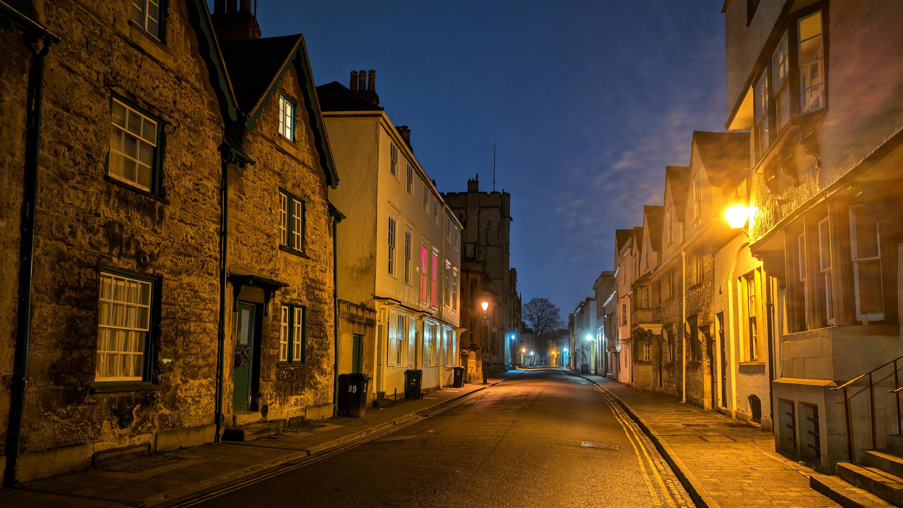 A night photo of an empty street in Oxford. There are street lights illuminating the scene. The street is lined with stone houses. 