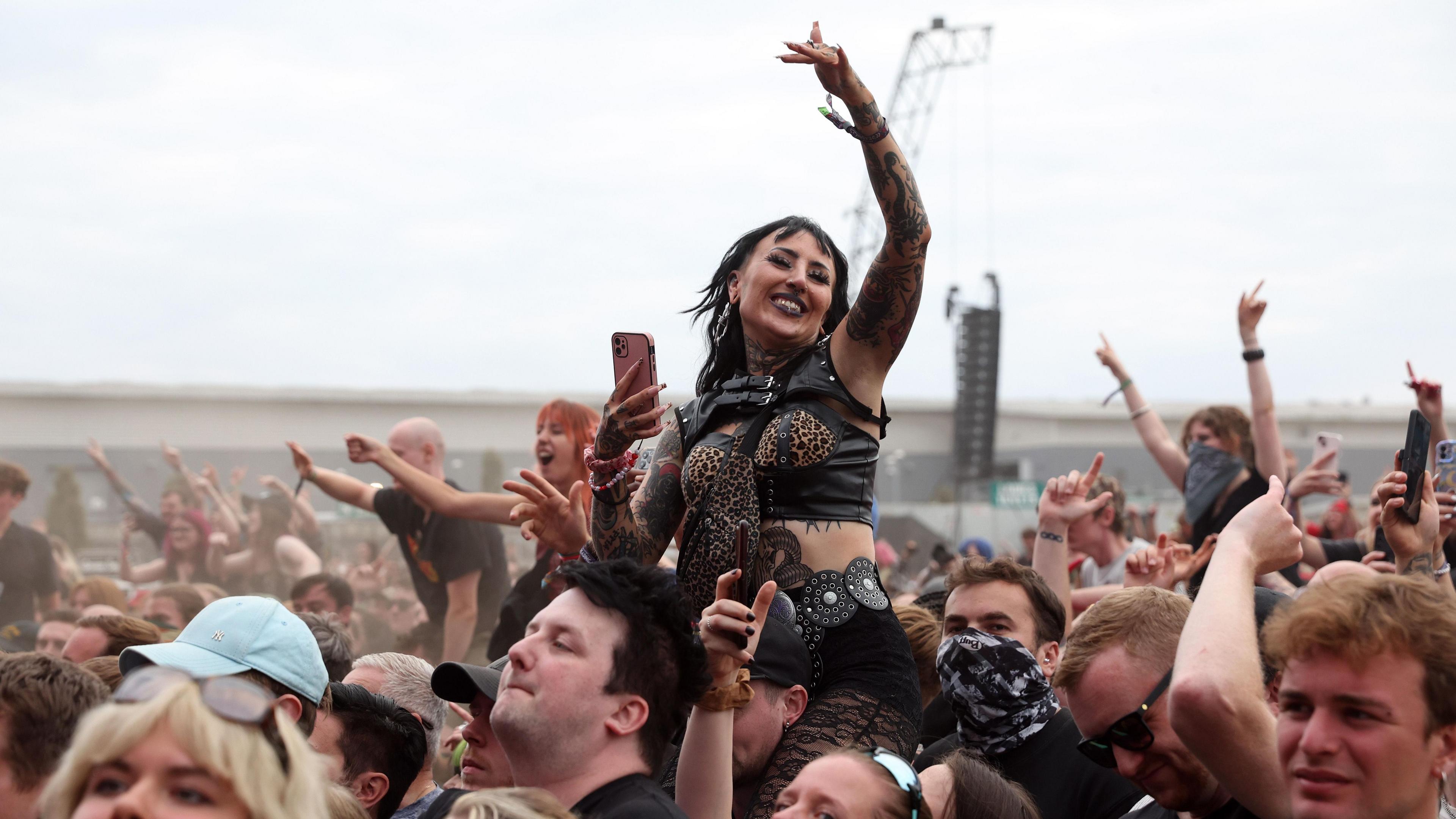 A fan on the shoulders of another soaks up the atmosphere, wearing all black and her hands in the air as Enter Shikari perform live onstage.