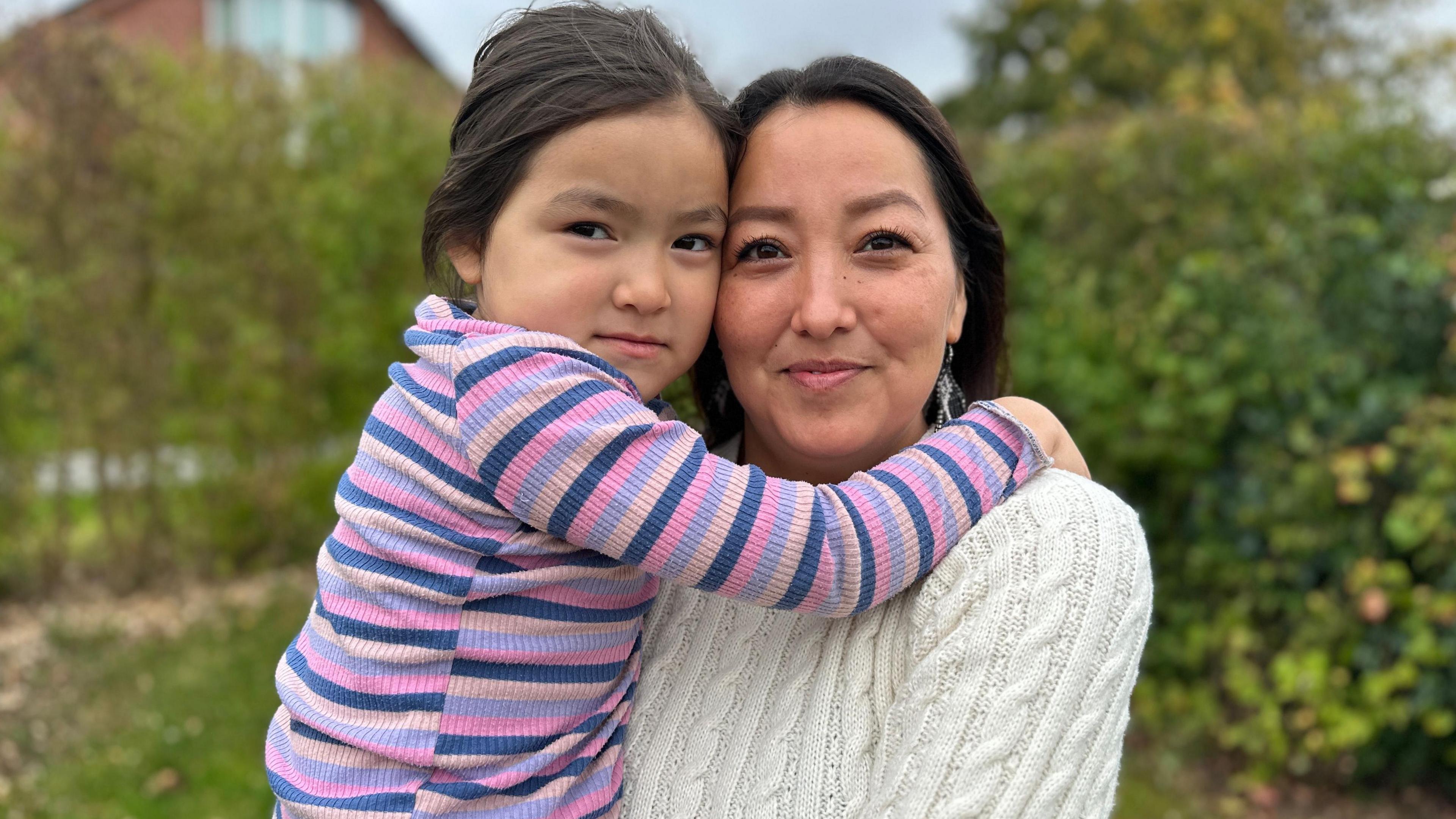Pilinguaq, who is dressed in a cream jumper with dangly earings, holds her daughter, who is wearing a striped pink, purple and blue top. The daughter is hugging her mother around her shoulders, while in the background there are blurred trees and bushes in front of a house.   