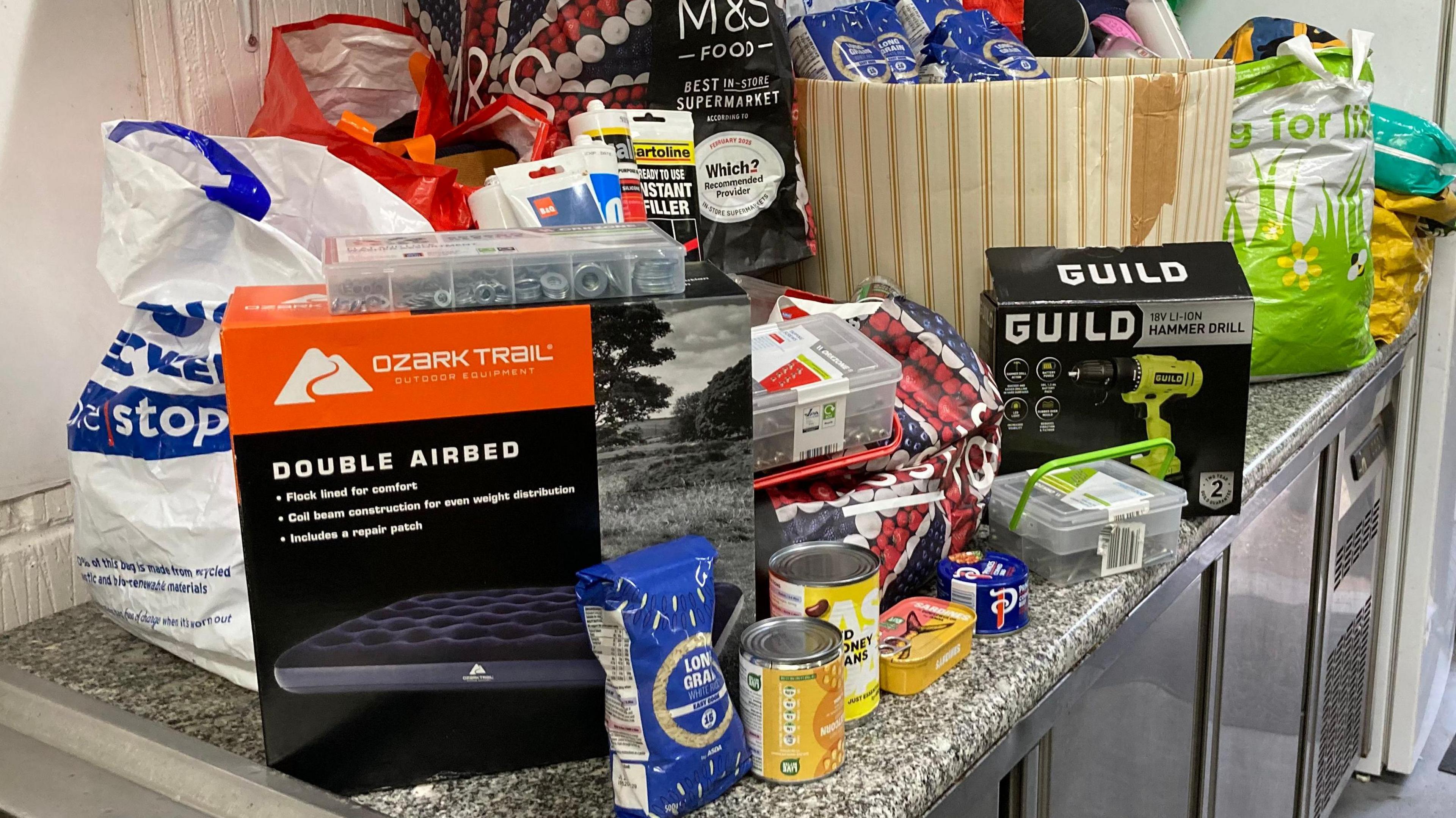 Tinned food, an inflatable mattress box, bags of rice and a hammer drill along with shopping bags full of items are on top of a grey kitchen worktop.