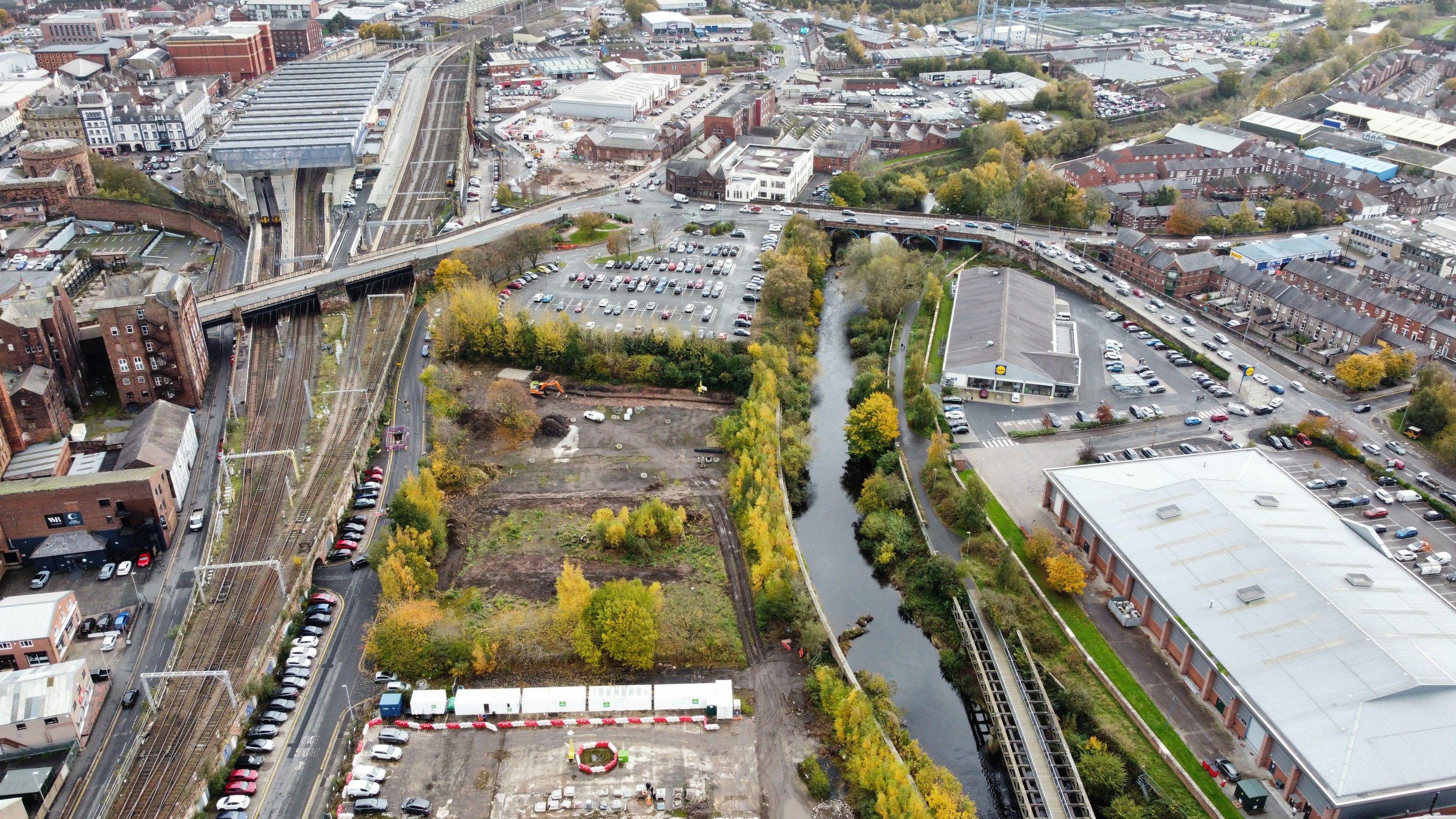 An aerial view of brownfield land with some trees and a car park in the distance. To the right runs a river and to the left runs a railway track. A bridge is in the distance and either side of the land is buildings and parked cars. 