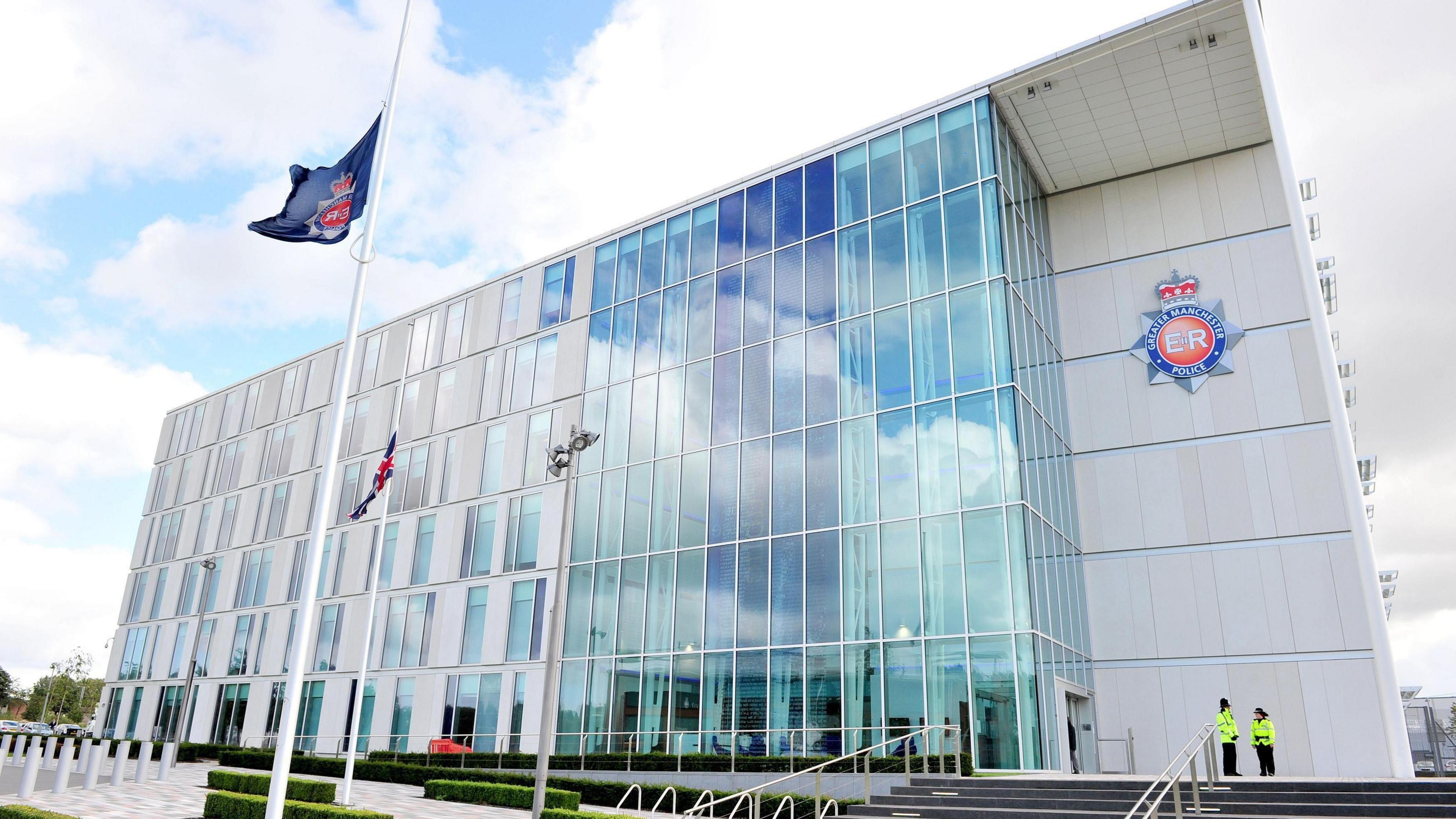 The grey, glass fronted Greater Manchester Police headquarters with the force's badge on the front of the building and two police officers standing at the top of steps that lead to the entrance doors