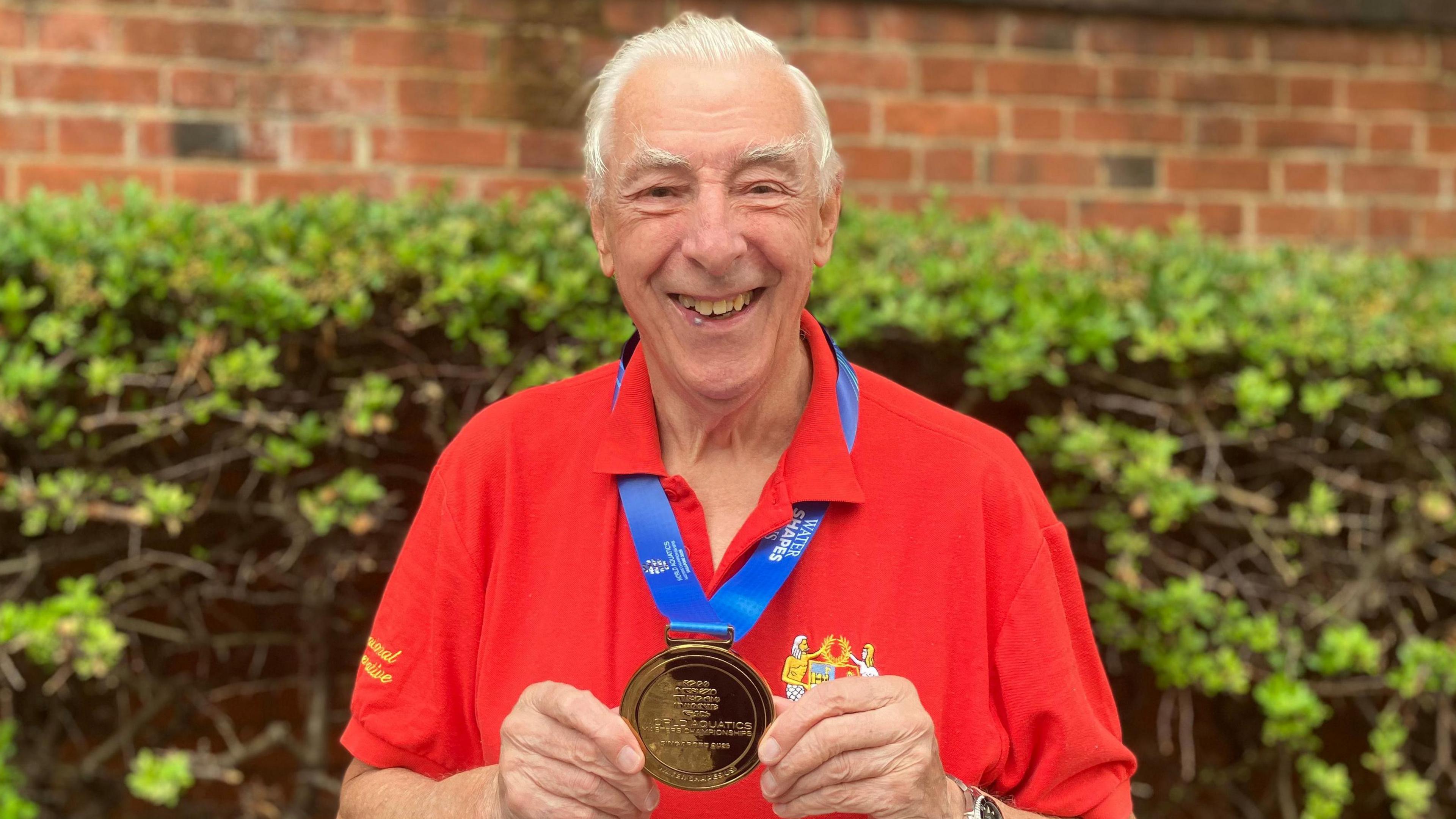 Michael Read smiles as he holds his gold medal that rests around his neck. He has short grey hair and wears a red polo T-shirt. His medal ribbon is a blue colour. He stands in front of a green bush and red brick wall.