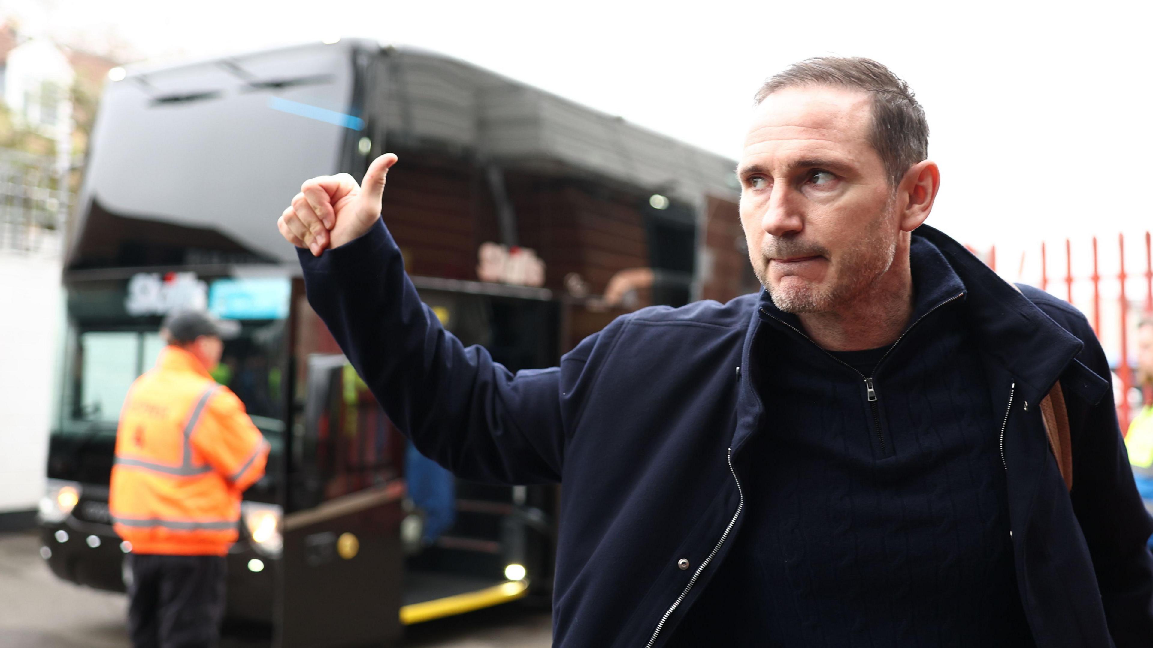 Frank Lampard gives a thumbs up in the direction of a bus outside a stadium.