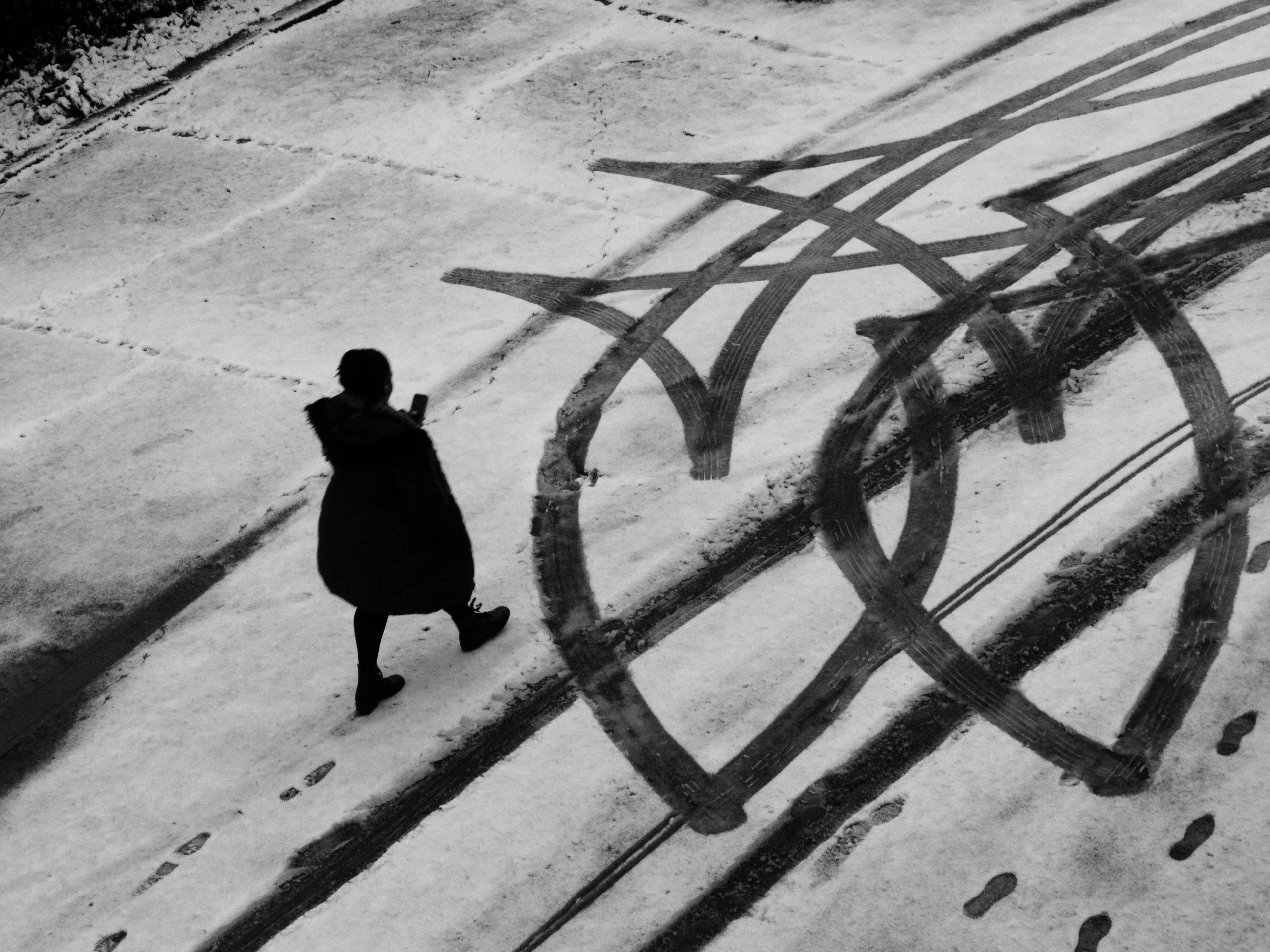 A woman walks on a snow-covered road with tyre tracks and footprints in front of her. The tyre tracks have made a pattern in the snow. The image is black and white.