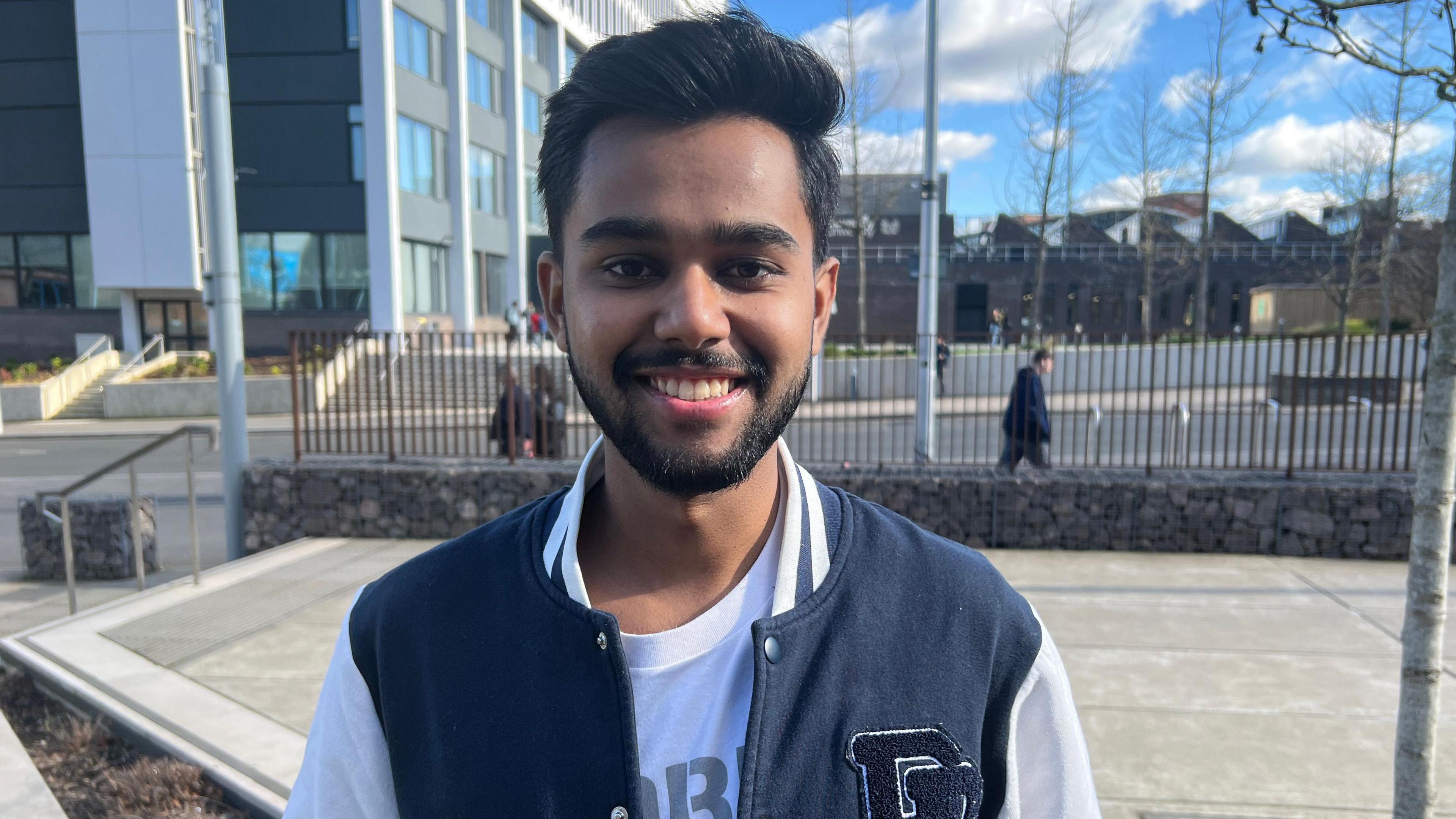 A young man with a beard and moustache smiles at the camera in a baseball jacket. He looks like he is standing on a university campus, with grey and brown buildings and railings behind him. People walk past behind the railings in the background. 