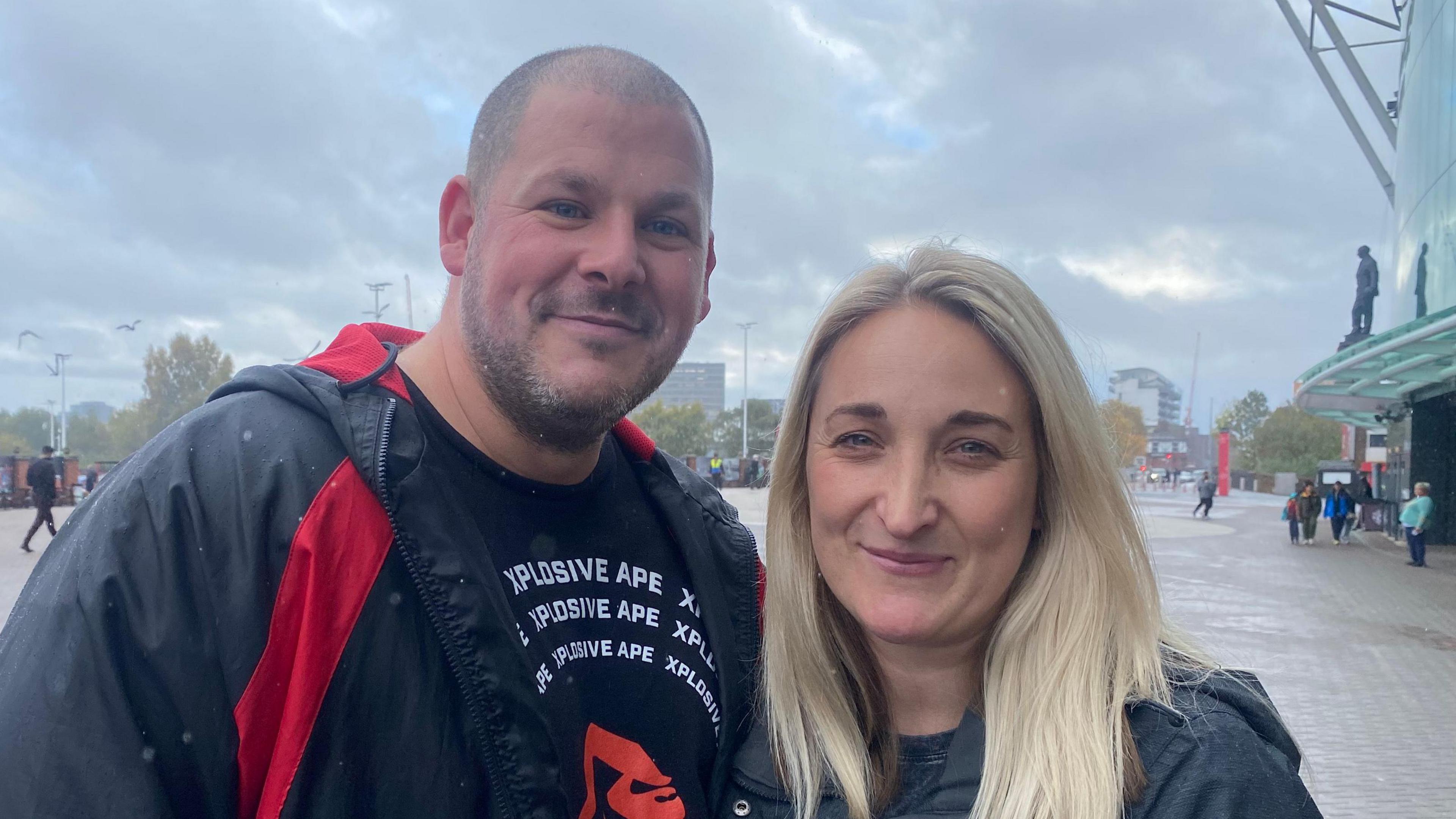 Chris and Carly White smile stood together outside Old Trafford on an overcast day.