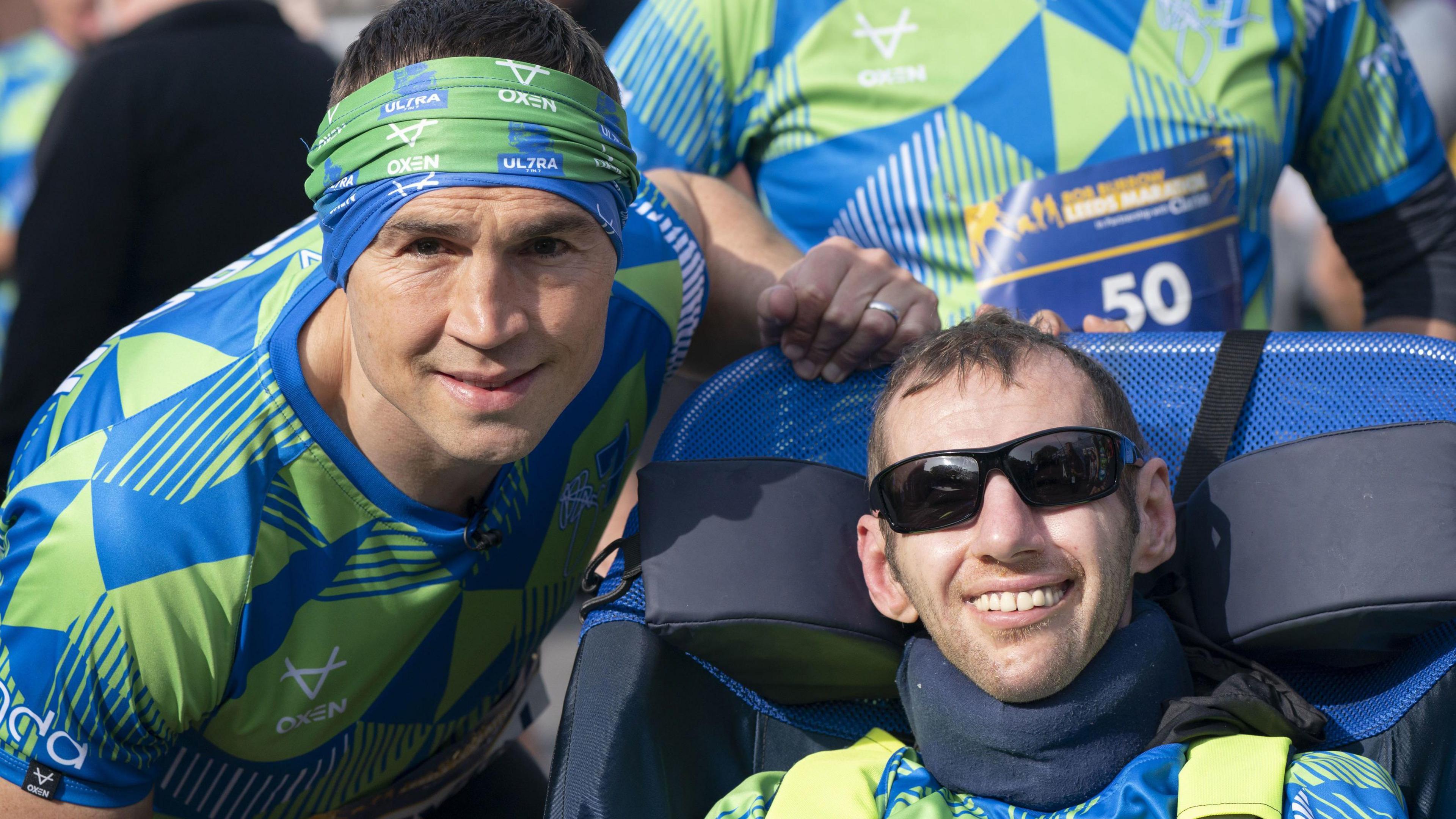 Dressed in blue and green Kevin Sinfield and Rob Burrow smile into the camera. Kevin is hunched slightly besides Rob's wheelchair. Rob is also wearing sunglasses. 