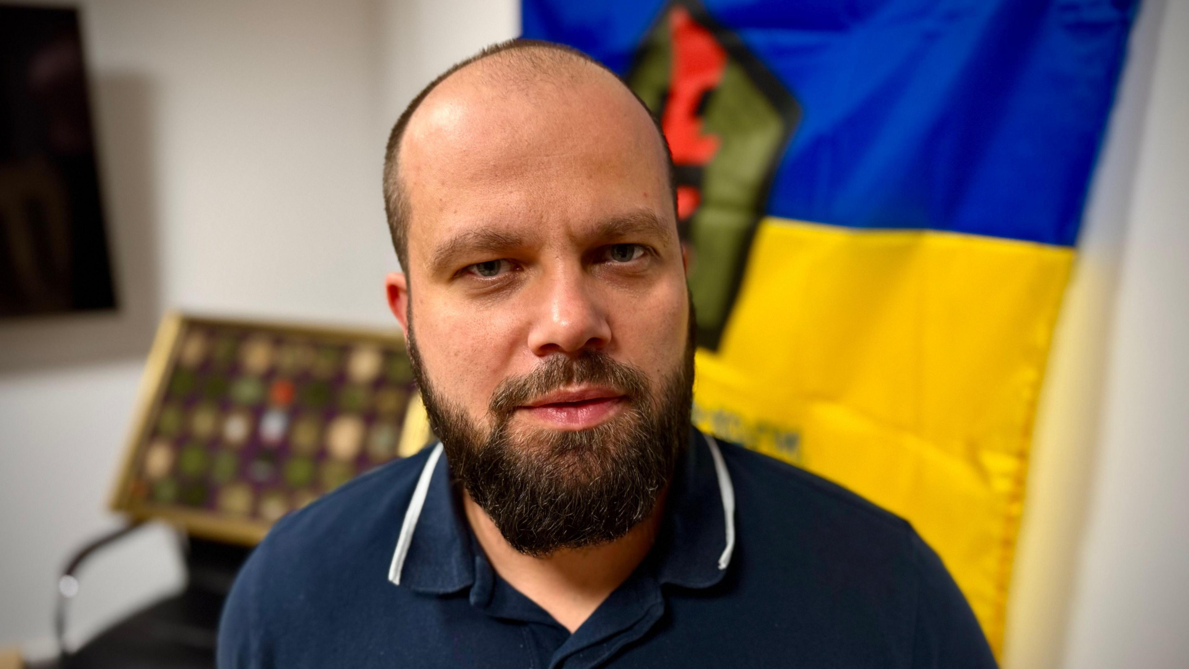 Oleg Lutsyk looks at the camera in a room where a Blue and yellow Ukraine flag is pinned to the wall behind him, a table and chair to the side. He is wearing a navy polo shirt trimmed in white.